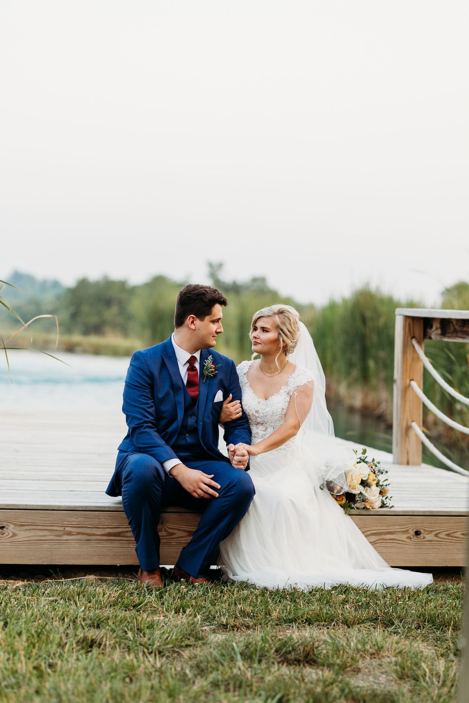 Couple shares romantic moment on wooden dock by lake, bride in white gown and groom in navy suit.