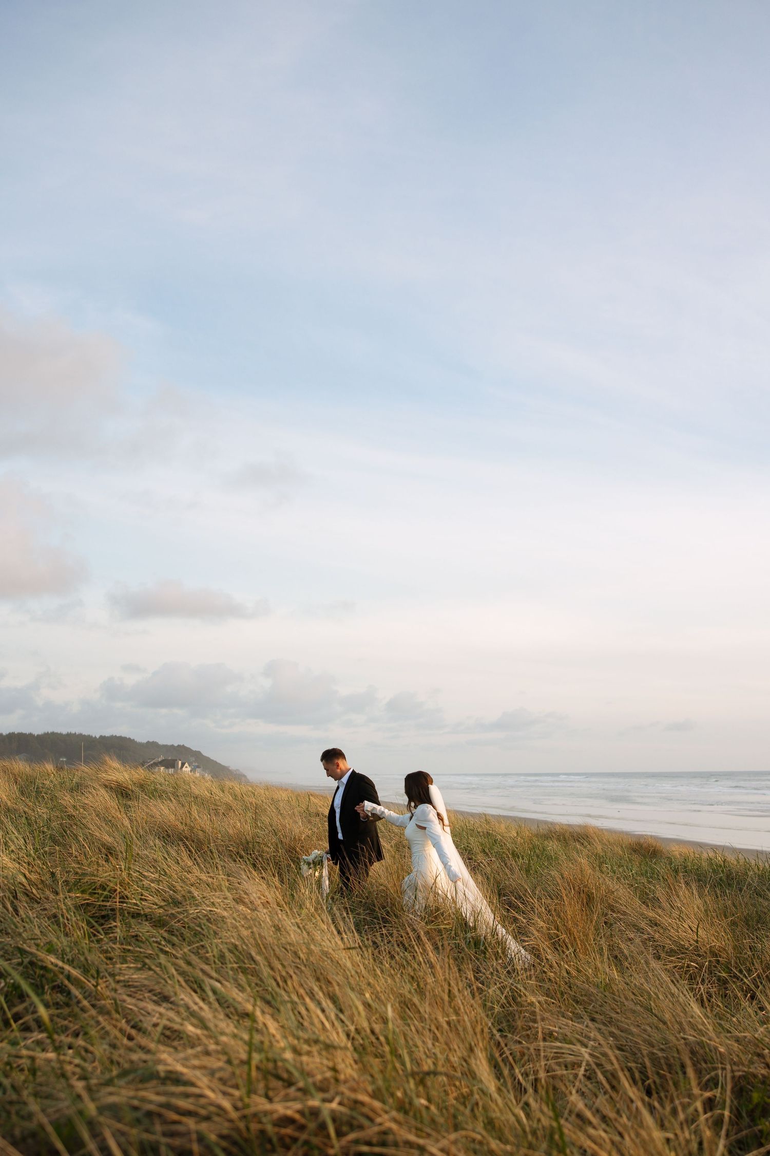 Couple walking through coastal dunes with ocean view at sunset.