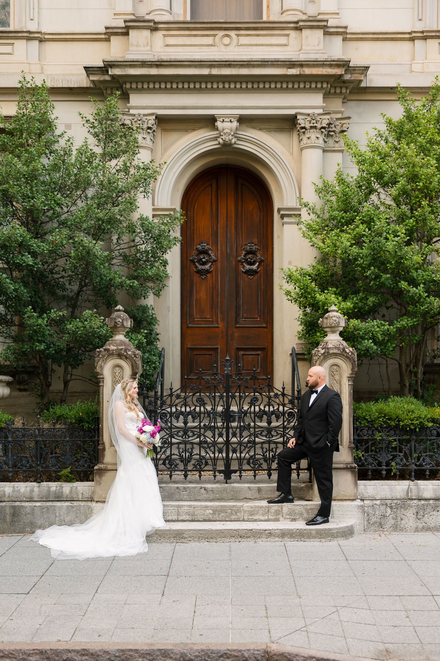 Bride and groom pose before ornate wooden door with iron gate at elegant stone building surrounded by lush greenery.
