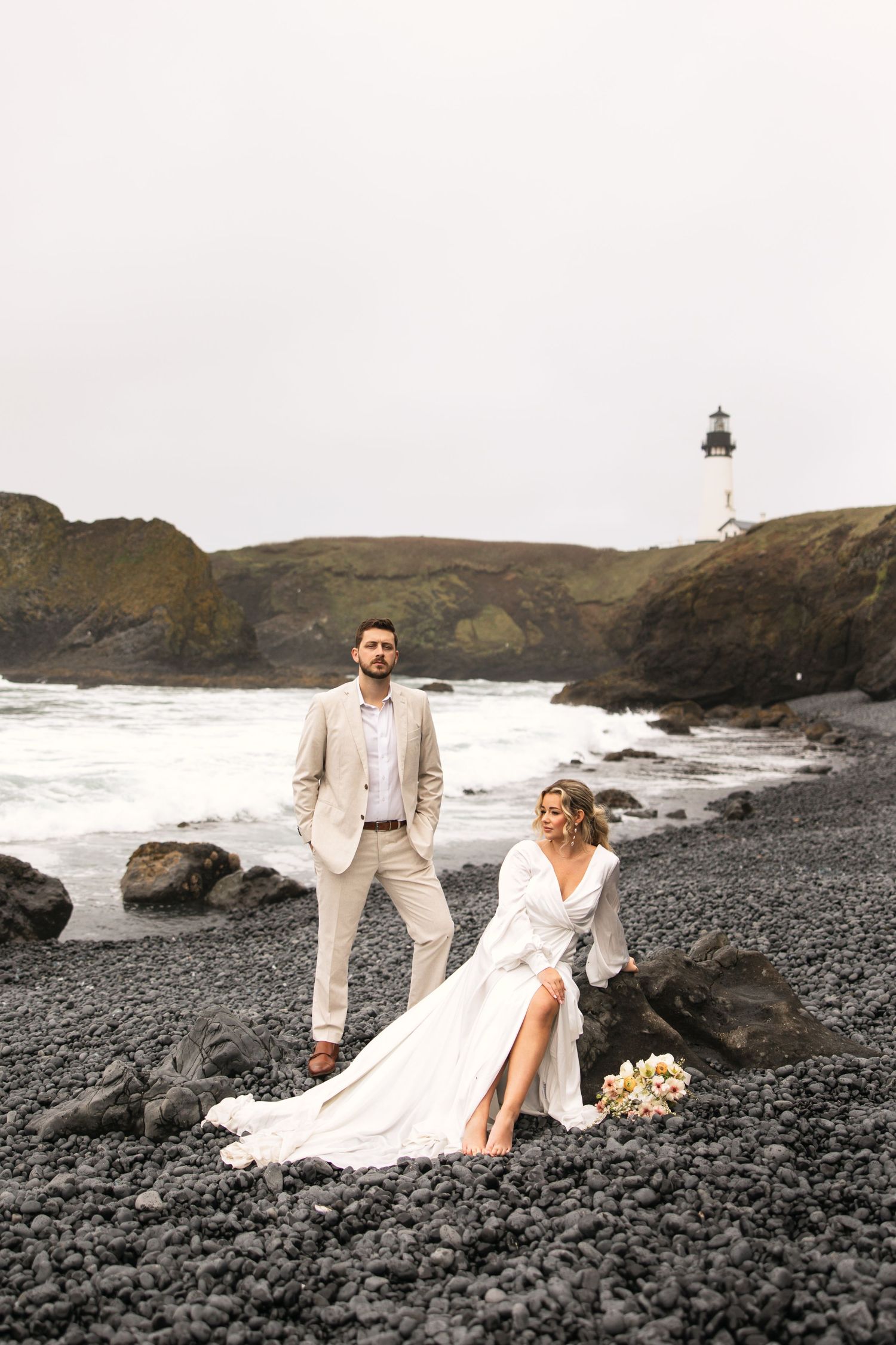 A seaside wedding portrait featuring a flowing white dress against black sand and rocky coastline.