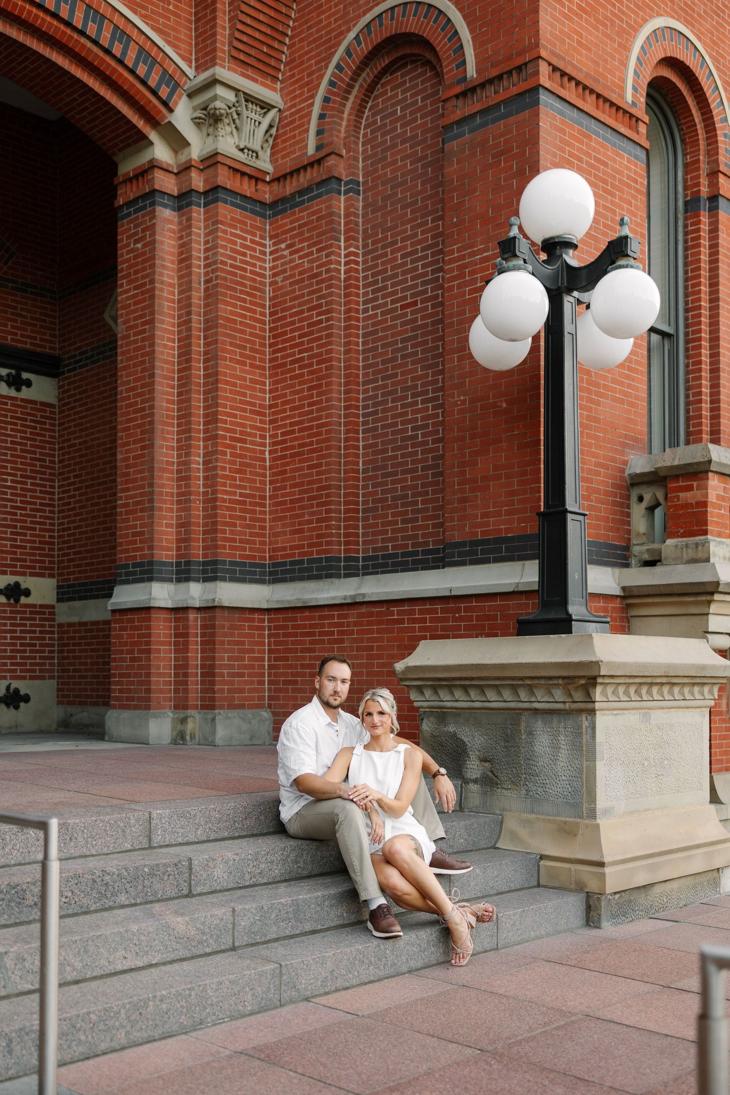 Couple in white outfits sit close together on gray stone steps beside a black lamppost in front of an ornate red brick building.