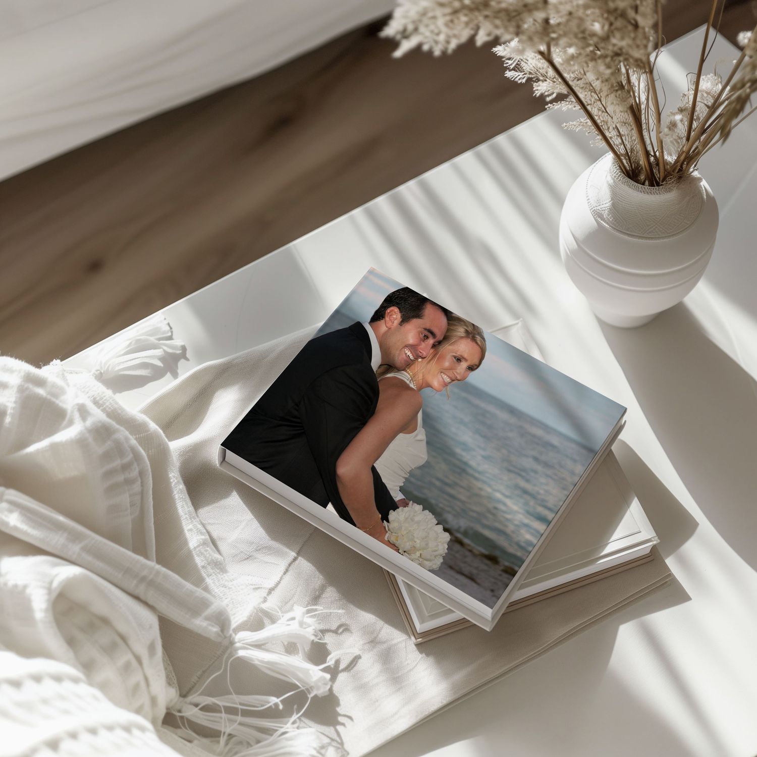 A wedding photo book with a couple's portrait cover displayed on a white table beside a vase with dried flowers.