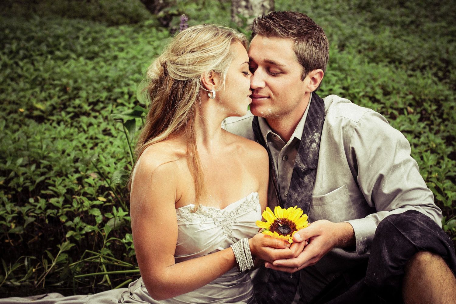 Romantic couple sharing a tender moment outdoors, holding a sunflower, dressed in wedding attire.