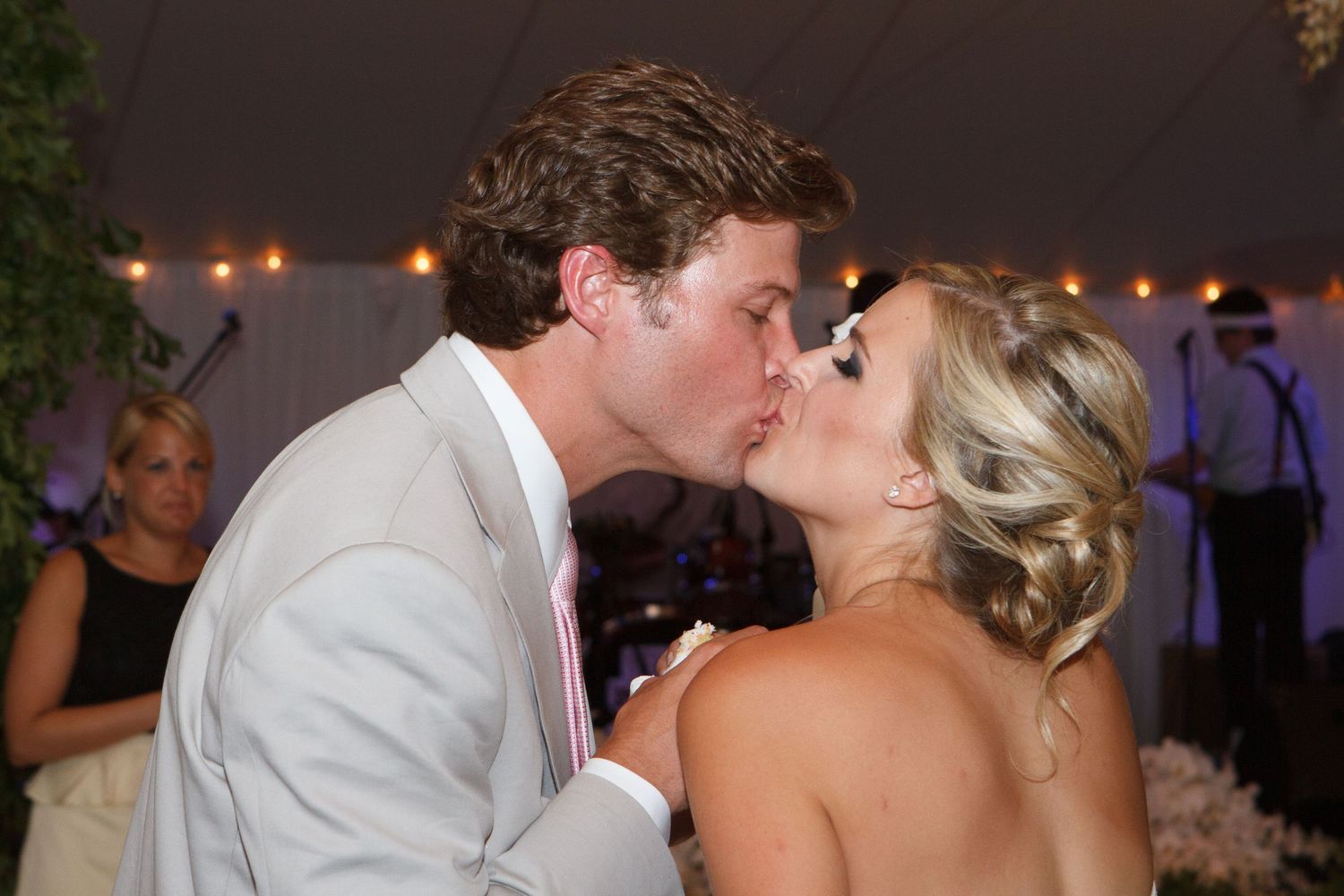 Couple sharing a romantic kiss at a wedding reception under a white tent with guests in the background.
