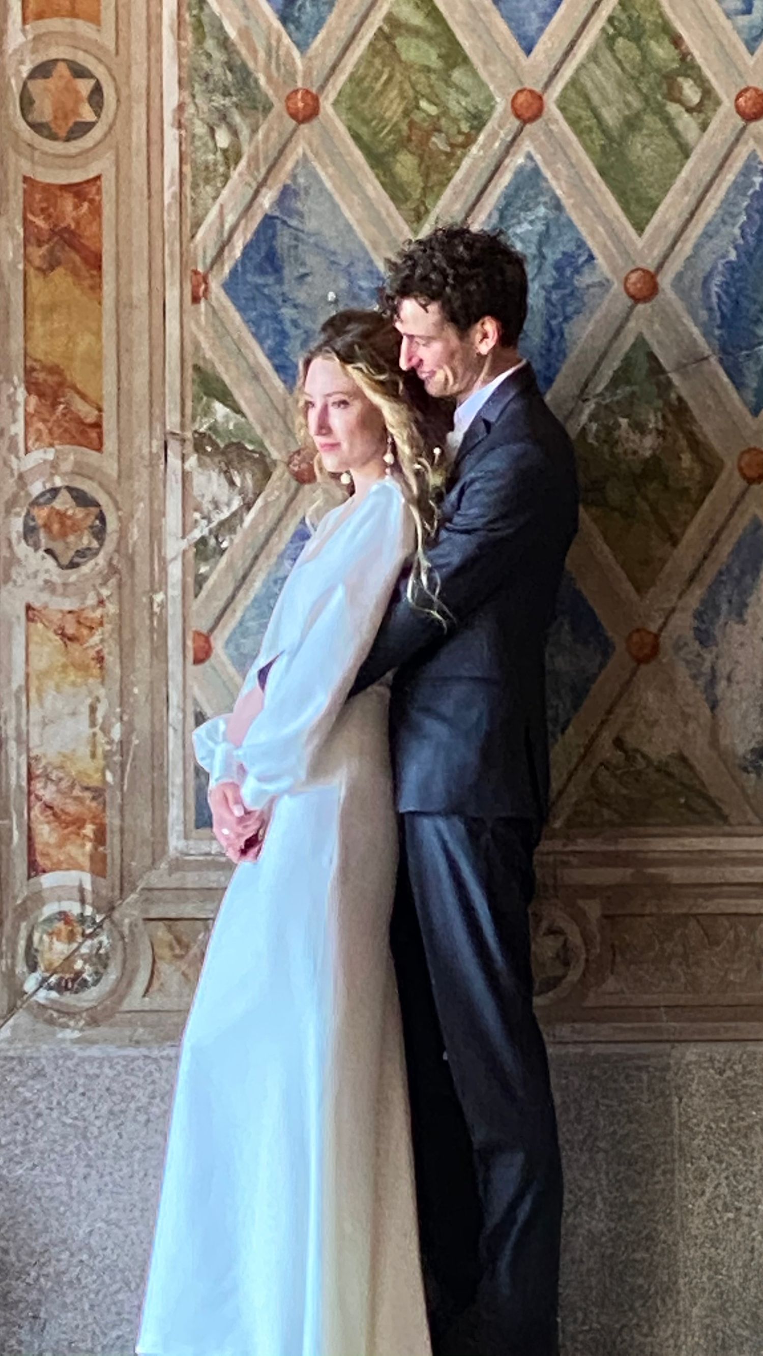 Elegant couple in wedding attire posing before ornate frescoed wall in historic Italian venue.