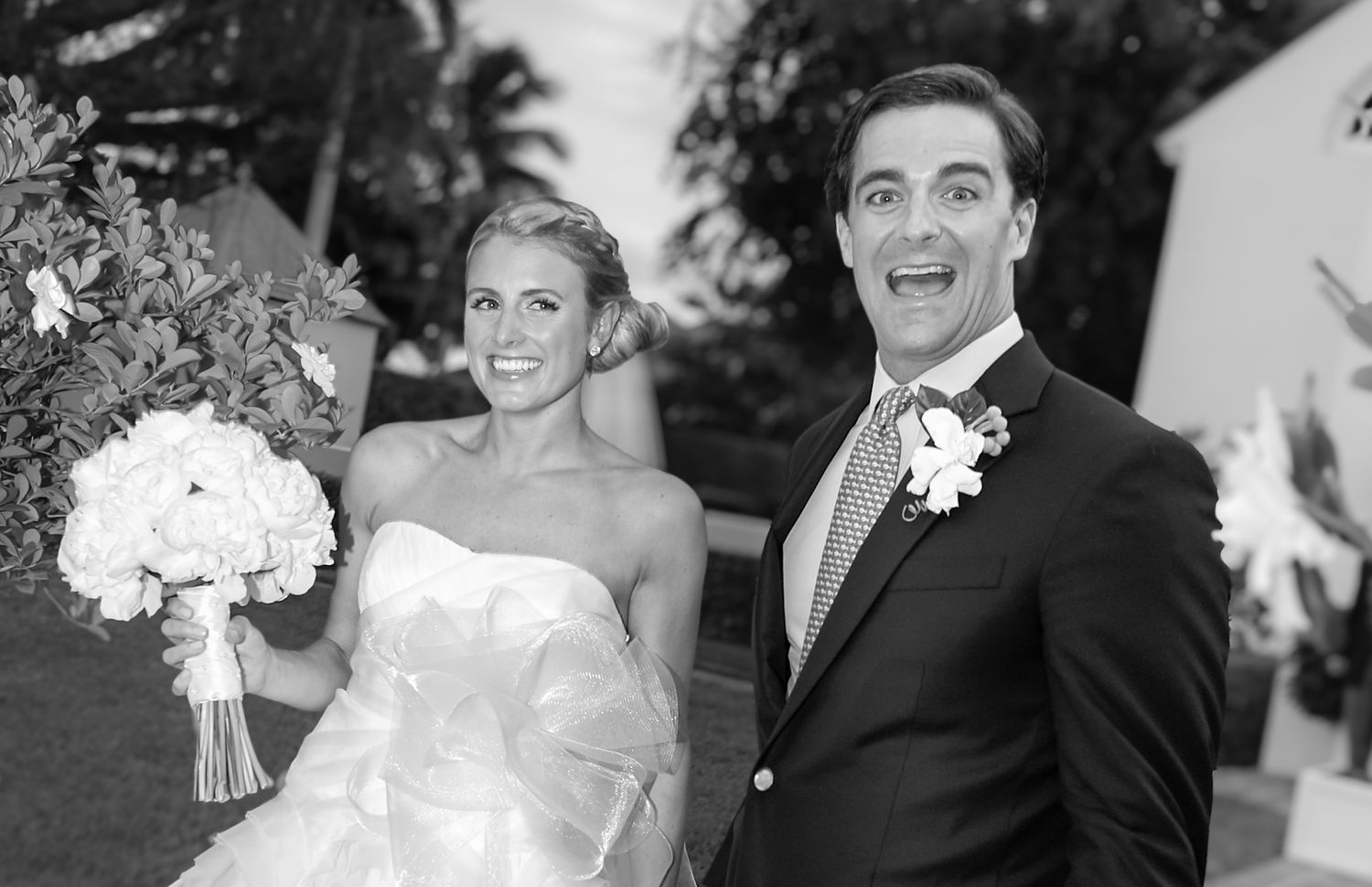 Black and white wedding photo of smiling bride holding bouquet and groom with excited expression outdoors.