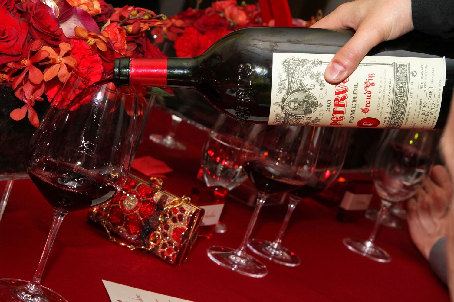 Hand pouring red wine from a bottle into glasses on a red table with floral arrangements at an elegant event.