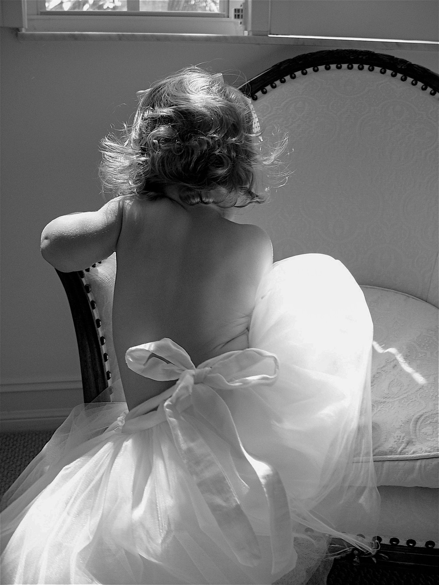 Black and white photo of a young girl in a backless white dress with bow, seated in a chair.