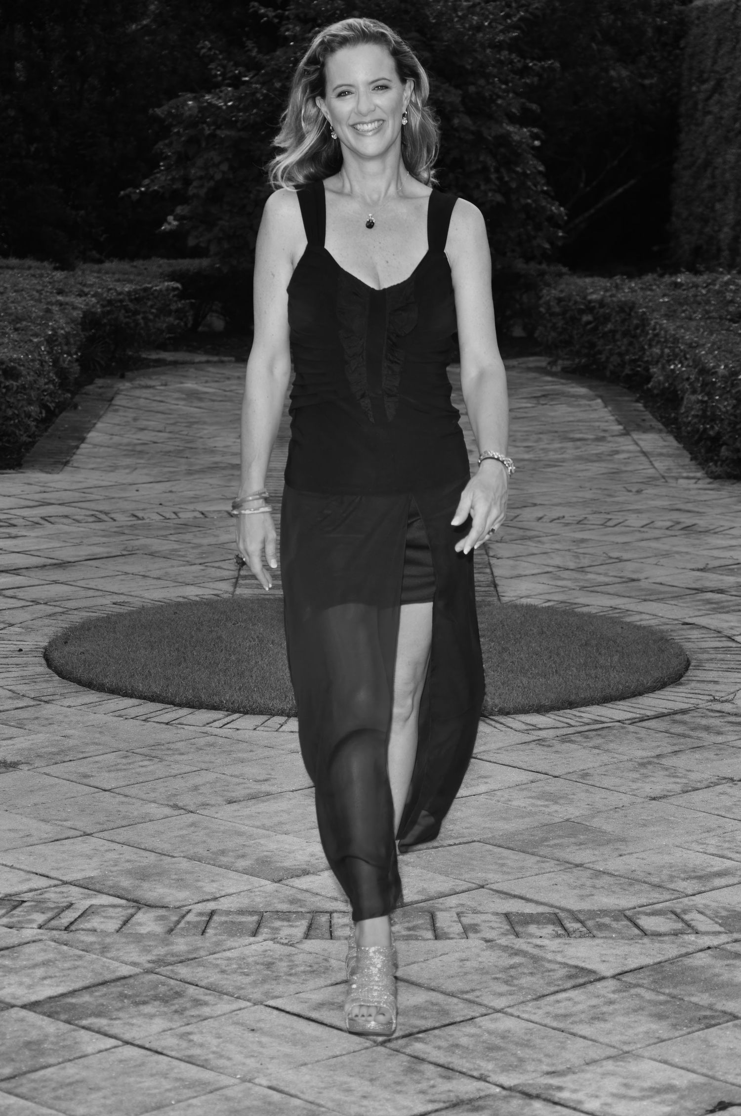 Woman in black tank top and skirt walks confidently along a stone garden path in a black and white photo.