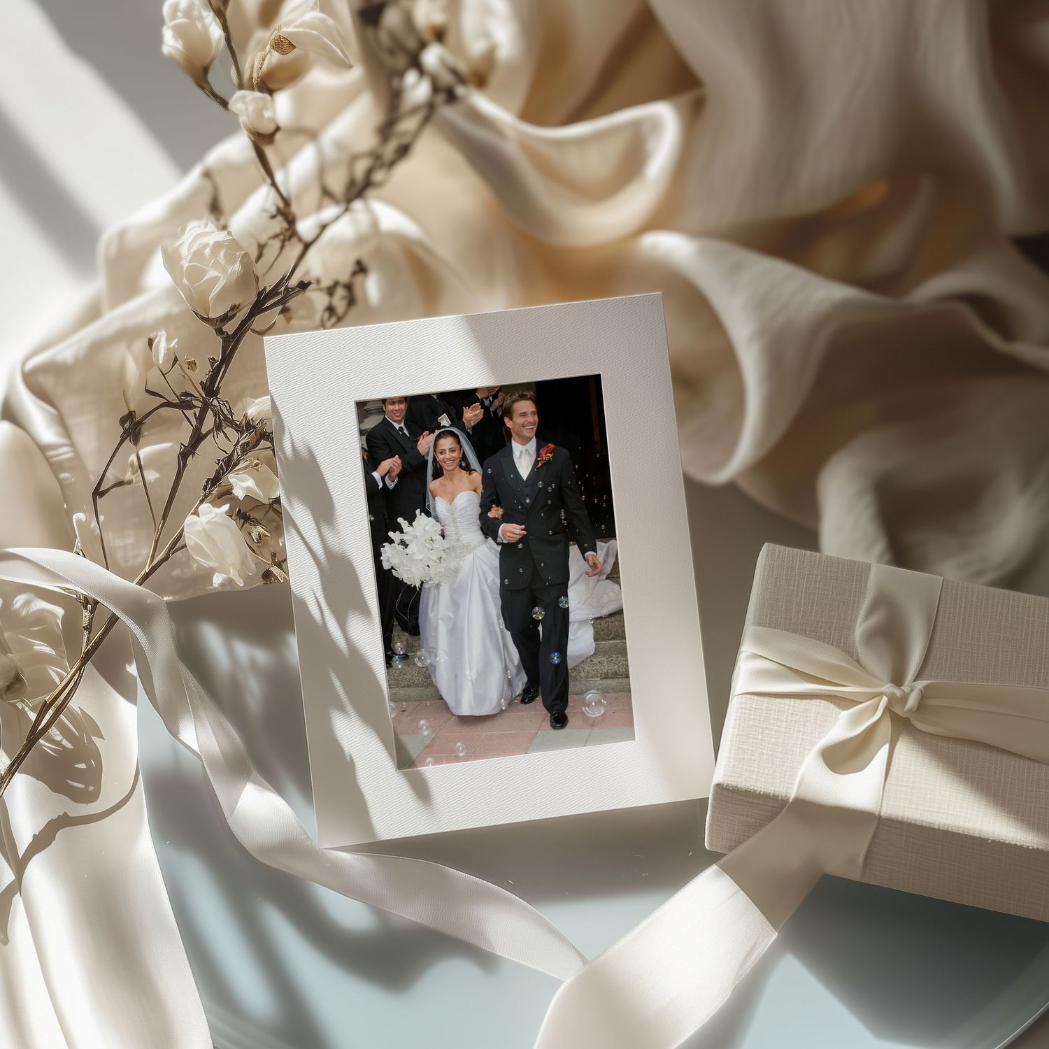 Elegant wedding photo in white frame surrounded by silk ribbon, gift box, and dried flowers on soft fabric.