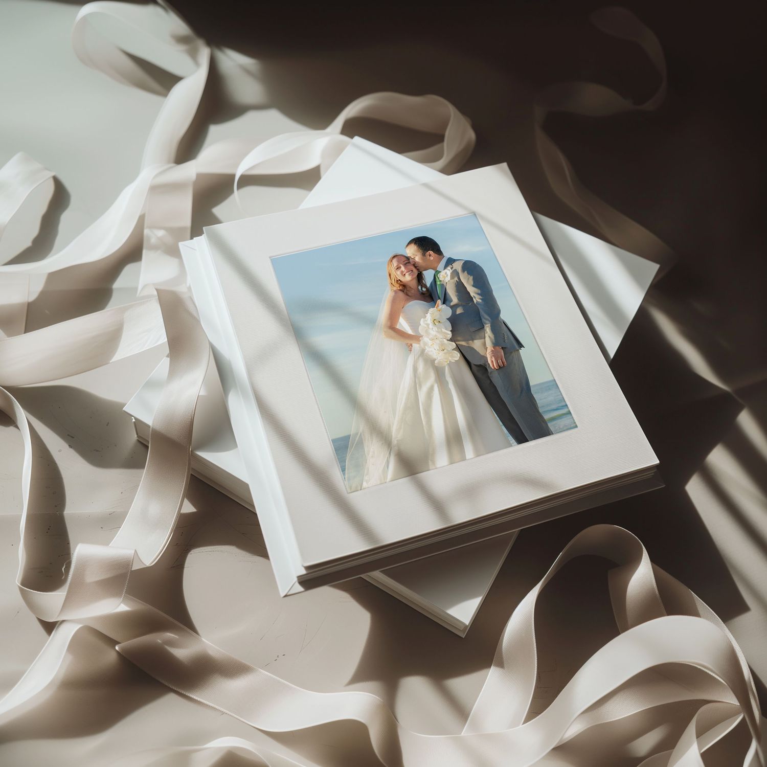 Elegant wedding photo book with a couple kissing on the cover, surrounded by soft satin ribbons in warm light.