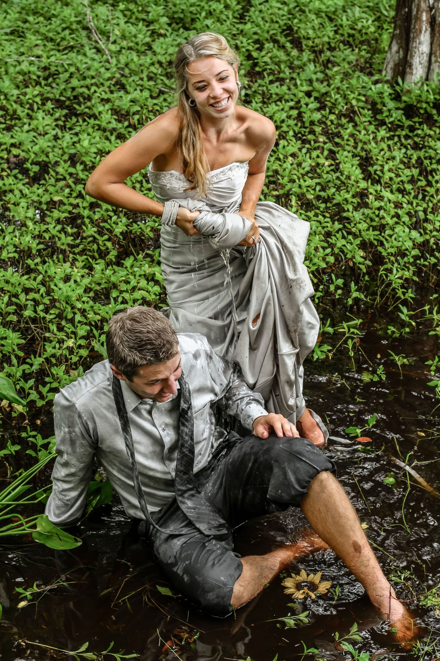 Bride in silver gown laughs as groom sits in muddy water during a fun trash-the-dress wedding photo shoot.