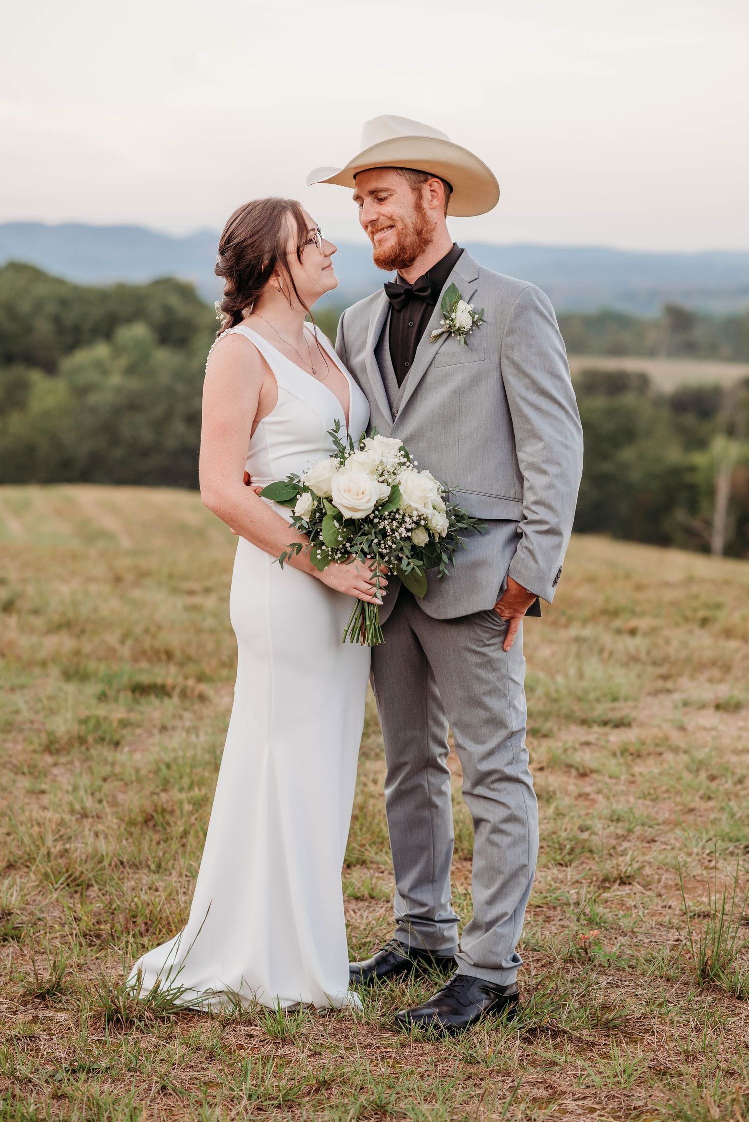 A couple in wedding attire stands together on a hilltop at sunset, with a white bouquet and rustic country style.