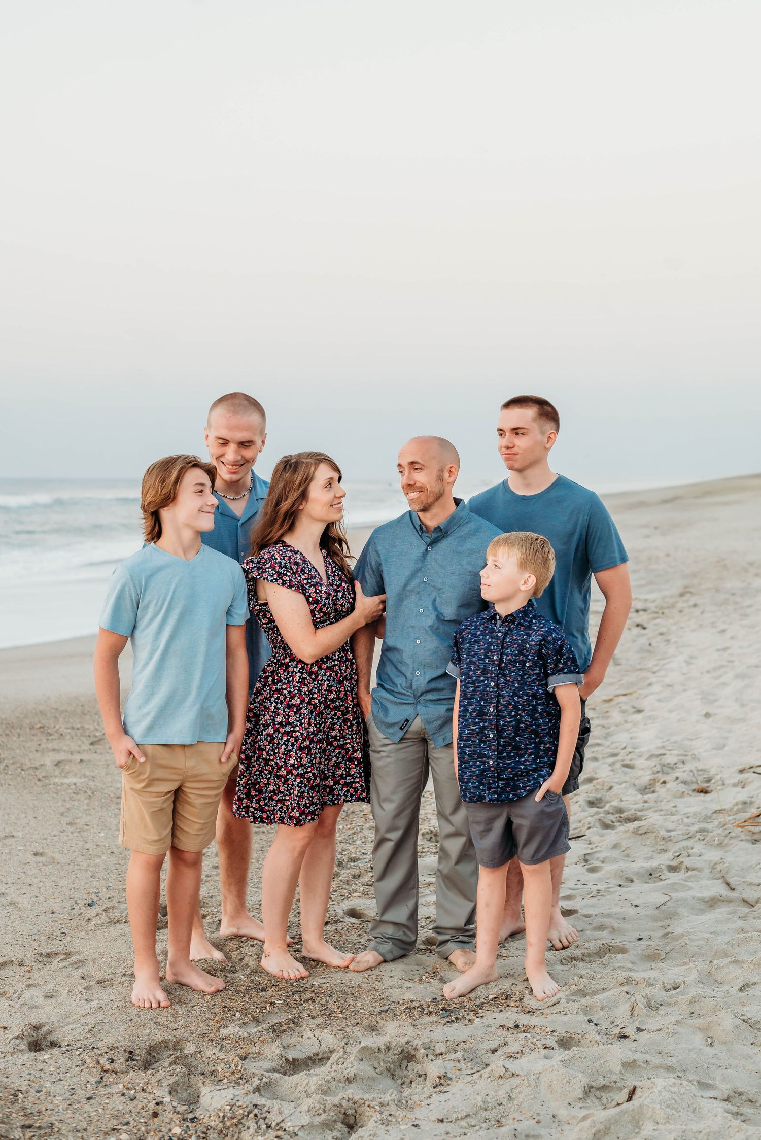 A group poses together on a beach at sunset wearing coordinated blue and navy outfits.
