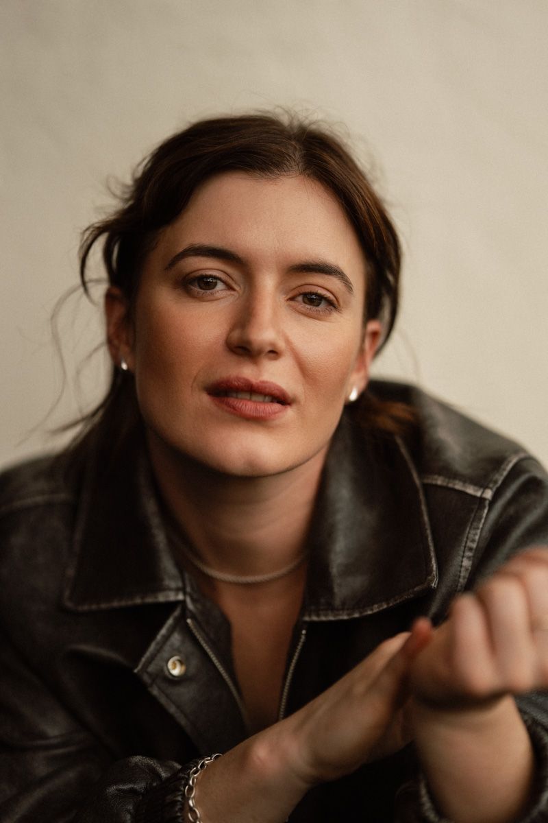 Young woman in black leather jacket with pearl earrings poses confidently against a light gray background.