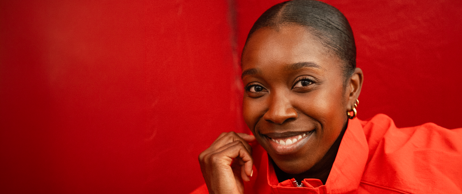 Smiling young Black woman in red outfit poses against vibrant red background with chin resting on hand.