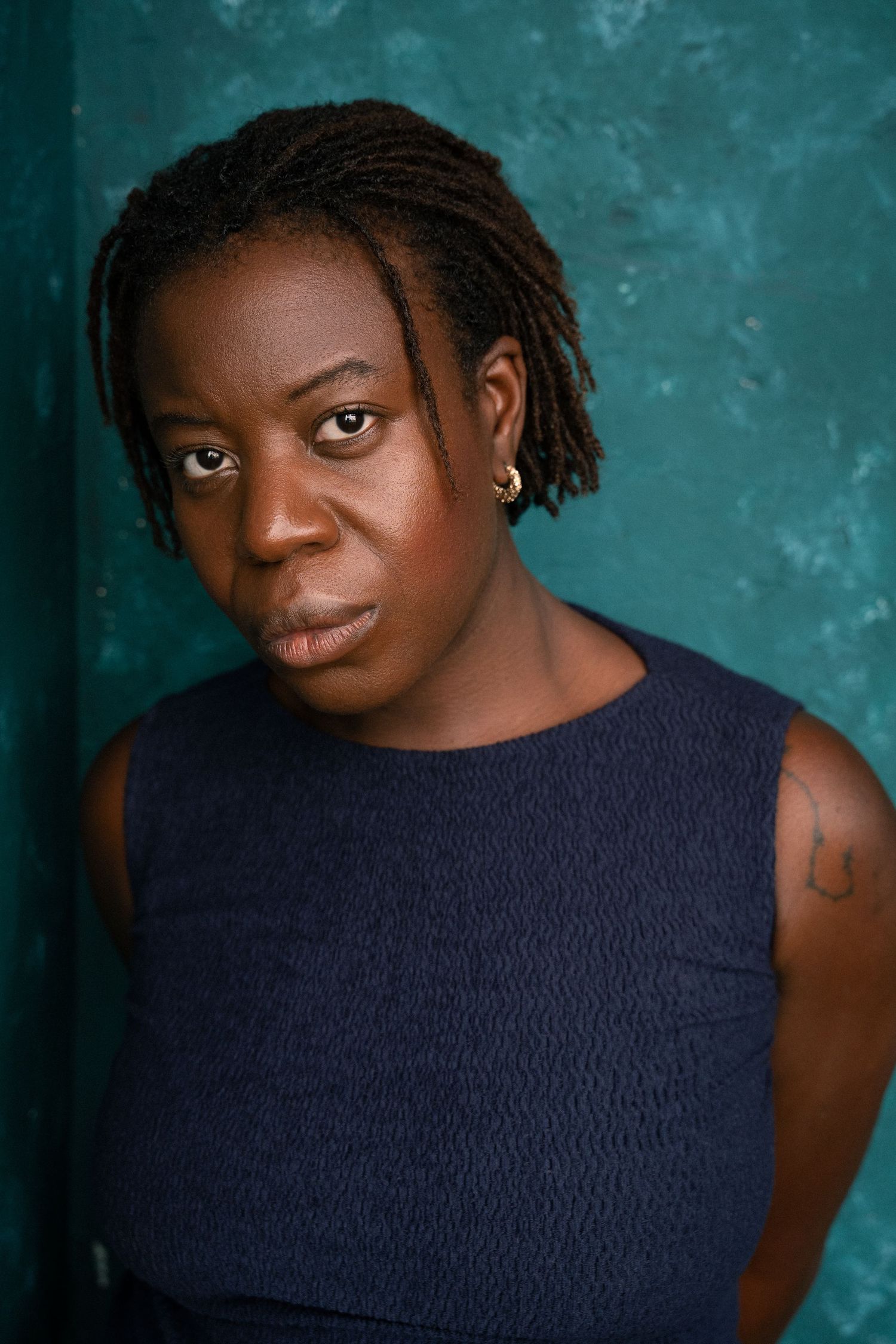 Woman with short dreadlocks in a navy sleeveless top against a teal textured background.