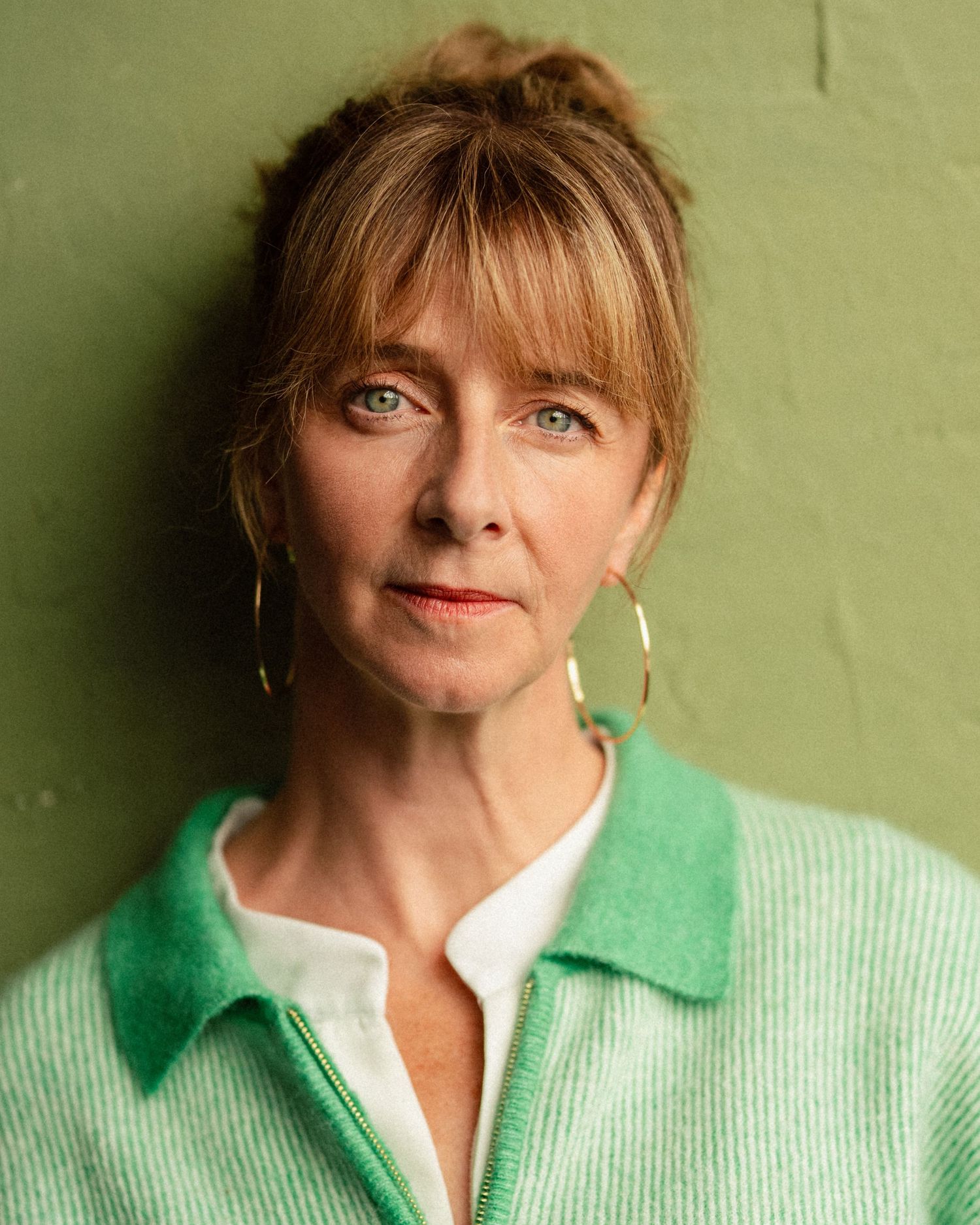 Middle-aged woman with blonde updo and bangs, wearing gold hoop earrings and green zip-up cardigan, posing against green wall.