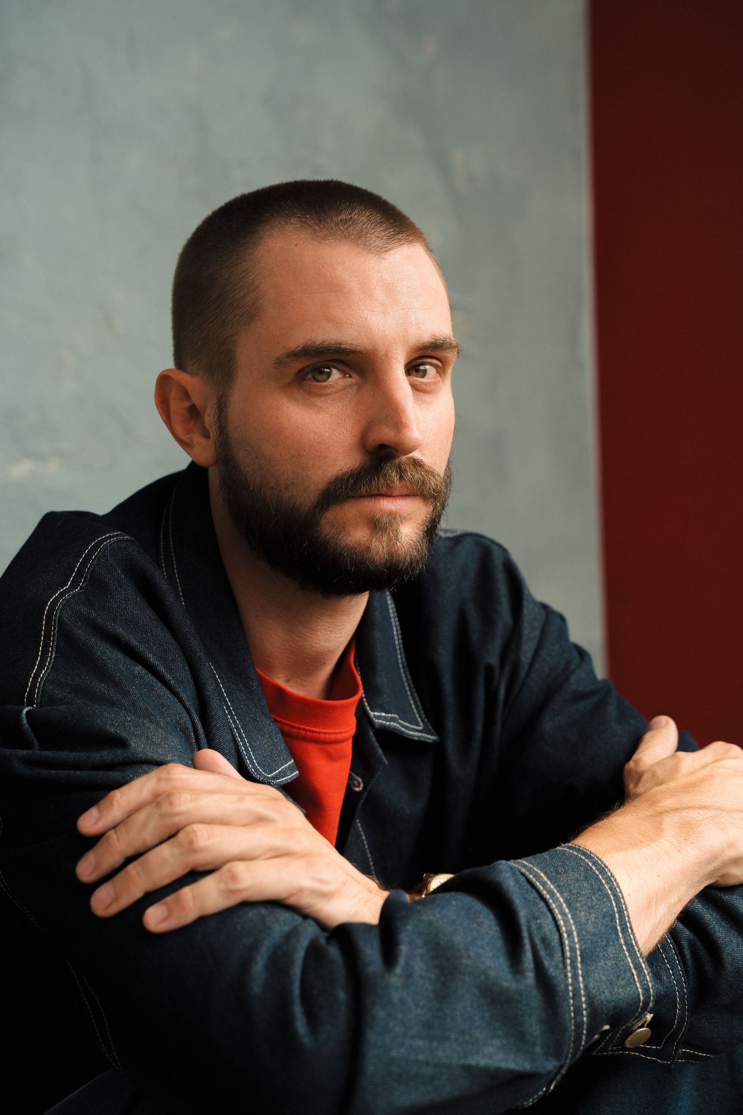 Bearded man with short hair in denim jacket sits with arms crossed, gazing thoughtfully against a gray wall.