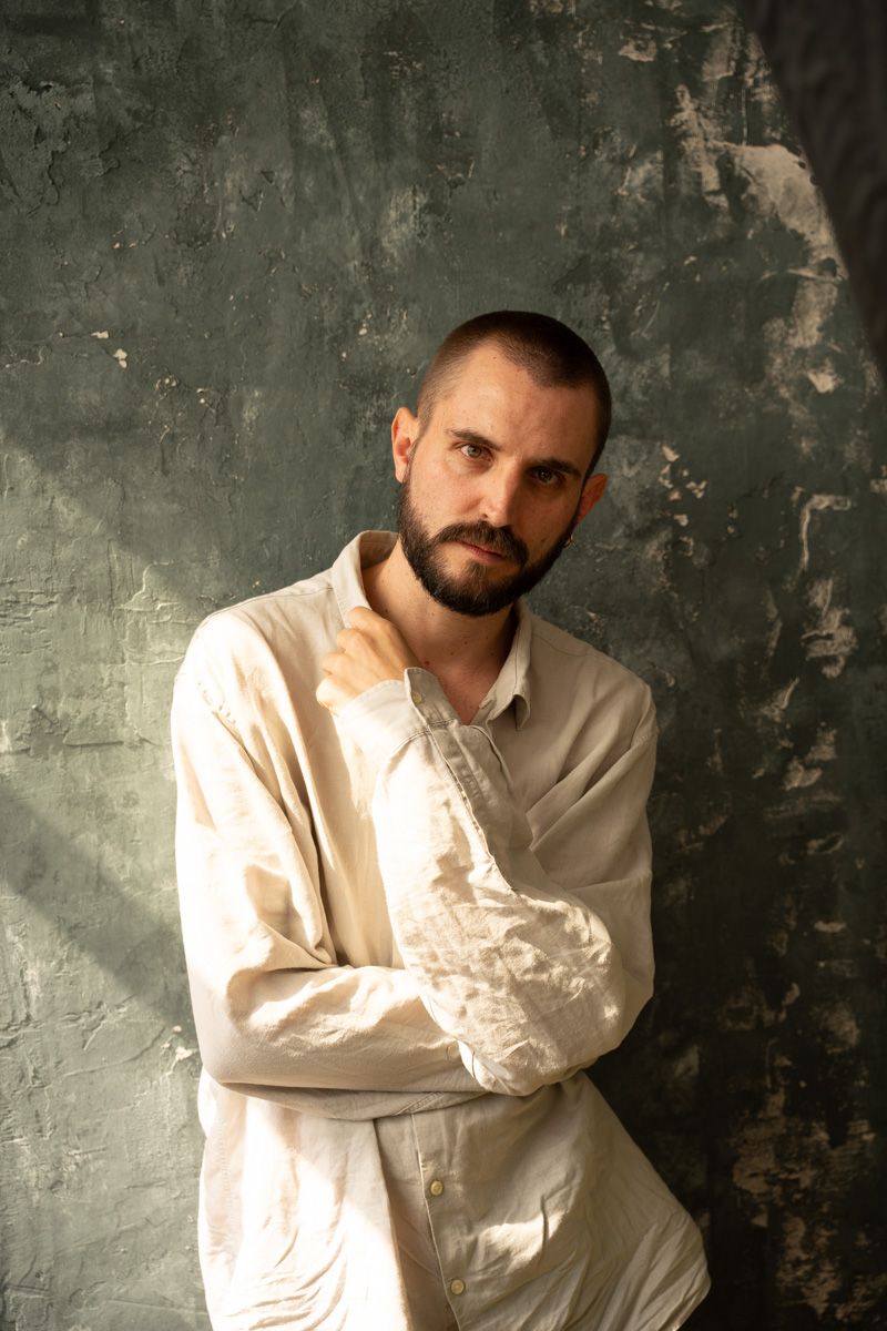 Bearded man with short hair in white linen shirt posing against textured gray wall in natural light.