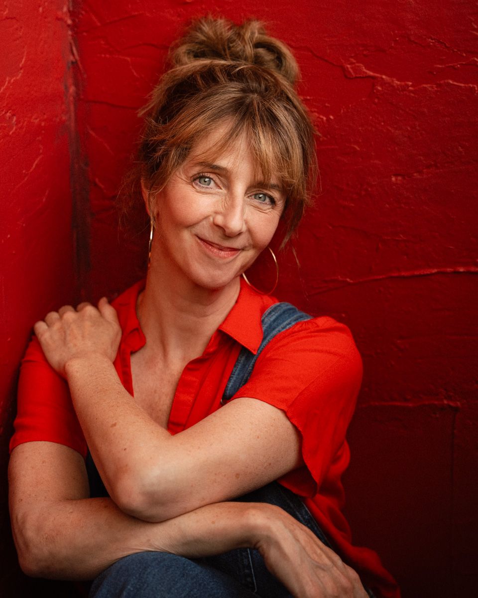 Woman in red shirt and denim overalls smiling against a red wall with arms crossed.