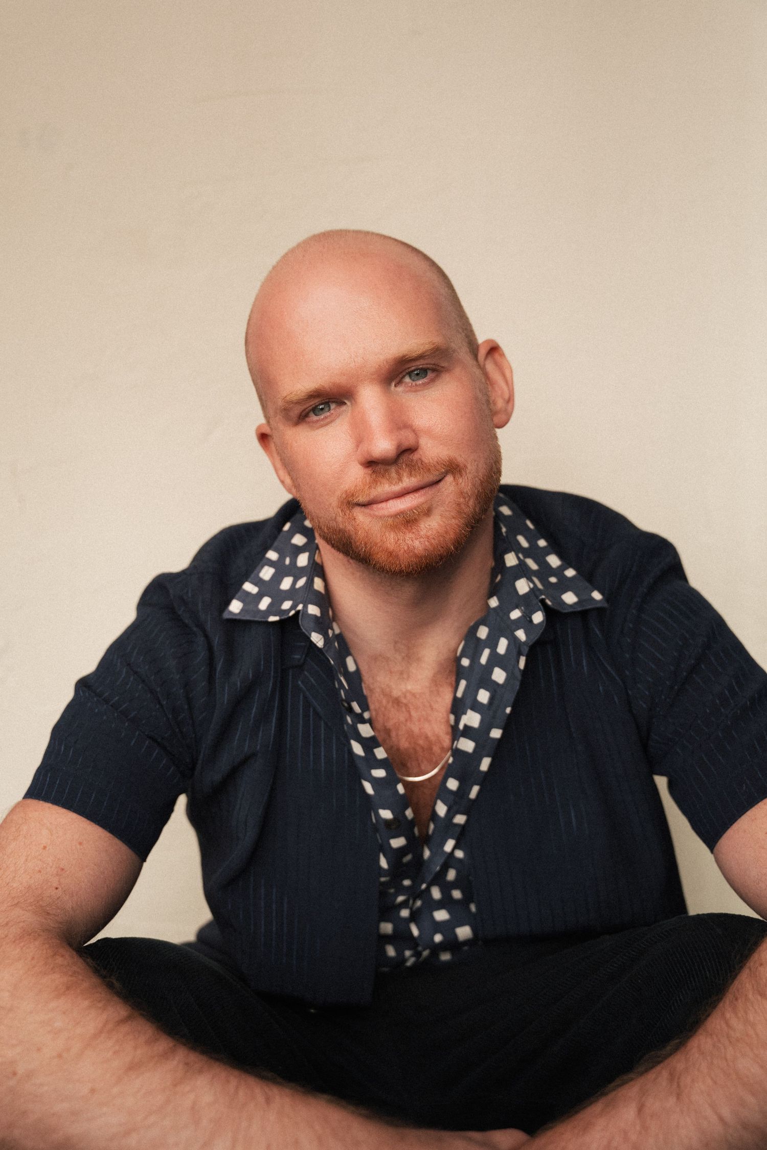 Bald man with red beard sits cross-legged, wearing navy blue shirt with patterned scarf, smiling at camera.