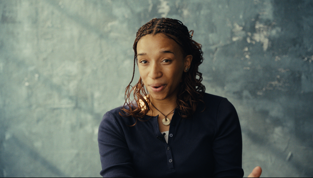 Young woman with braided hair in navy cardigan gesturing expressively against textured gray background.
