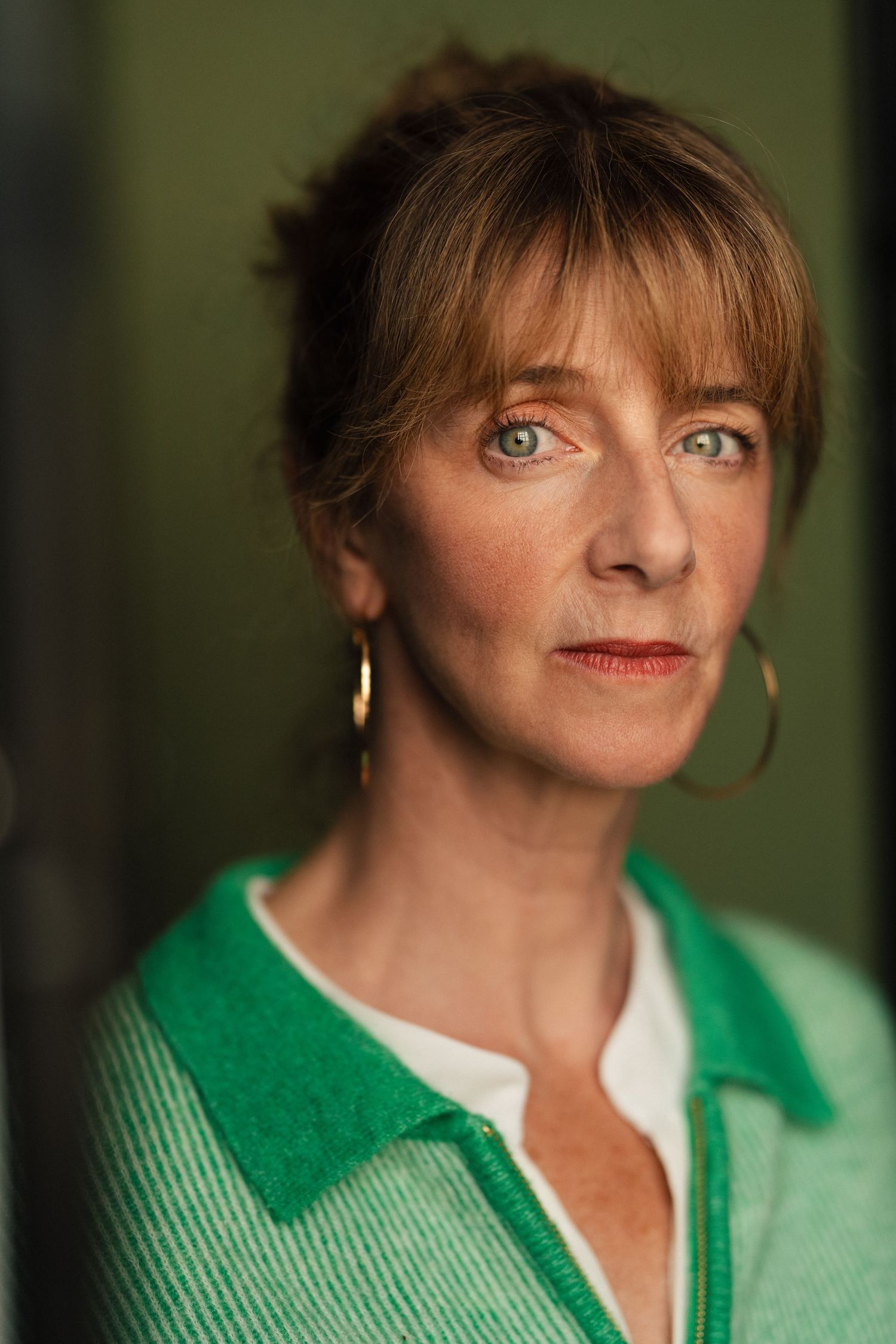 A middle-aged woman with green eyes and auburn hair wearing gold hoop earrings and a green cardigan poses for a portrait.