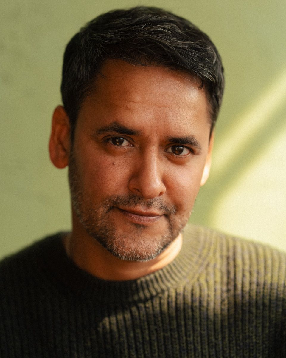 Middle-aged man with short dark hair and stubble wearing a dark sweater, photographed against a green background.