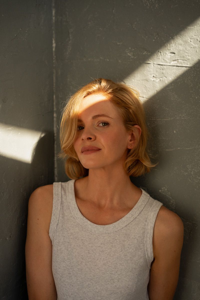 Young blonde woman in gray tank top leaning against gray wall with sunlight casting dramatic shadows.
