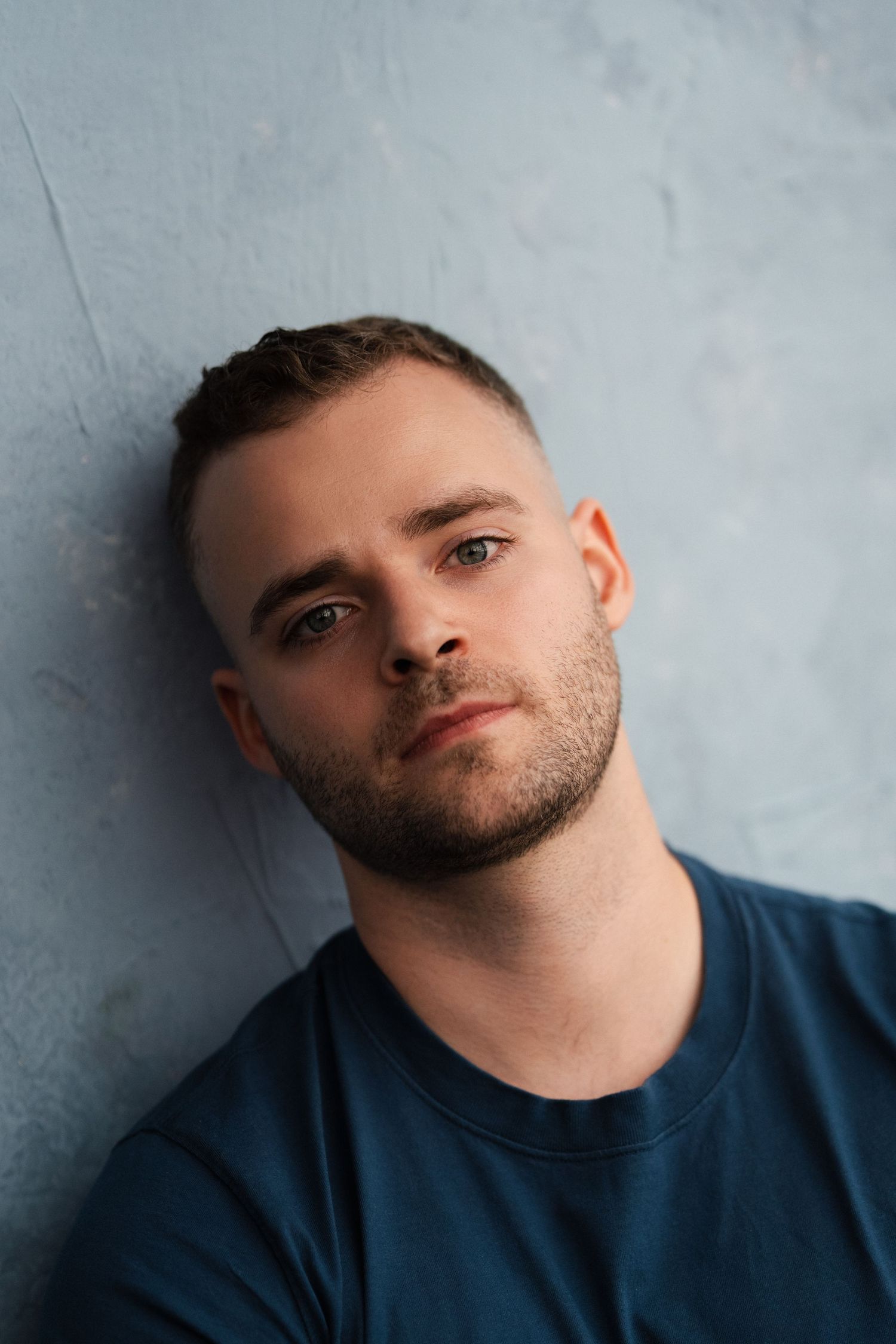 Young man with short hair and beard in blue t-shirt leaning against gray wall, looking at camera.