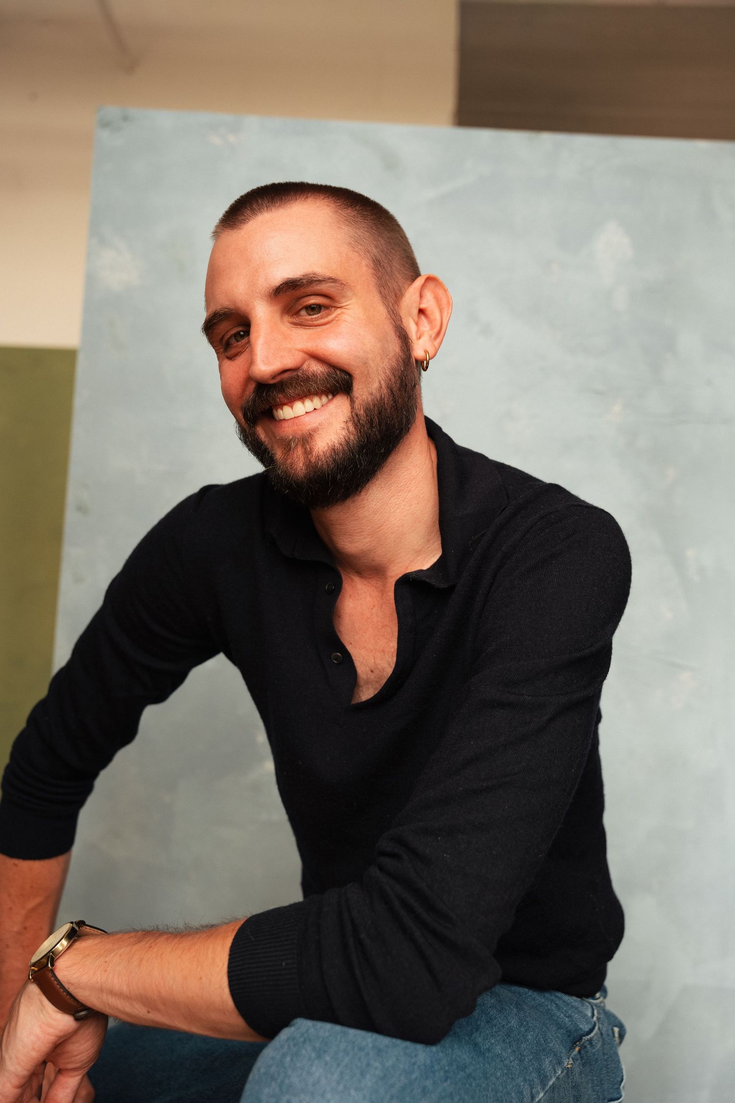 Smiling bearded man with short hair wearing a black shirt and jeans, sitting relaxed against a blue wall.