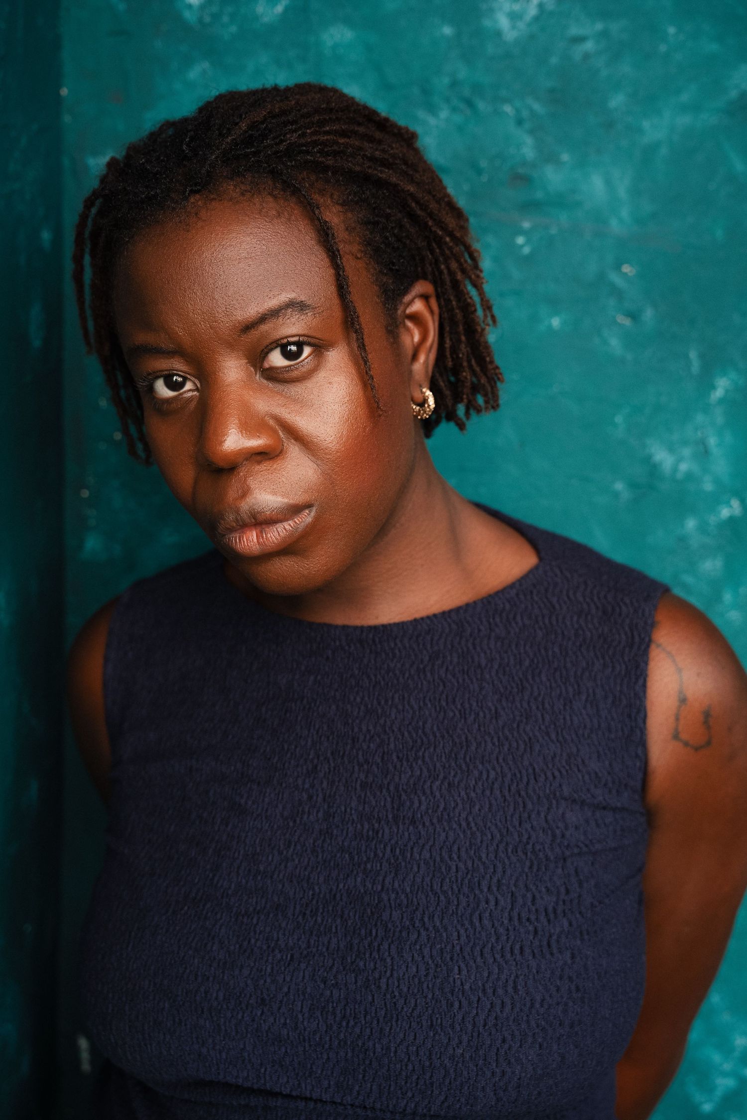 A woman with short dreadlocks wearing a navy sleeveless top poses against a teal wall.