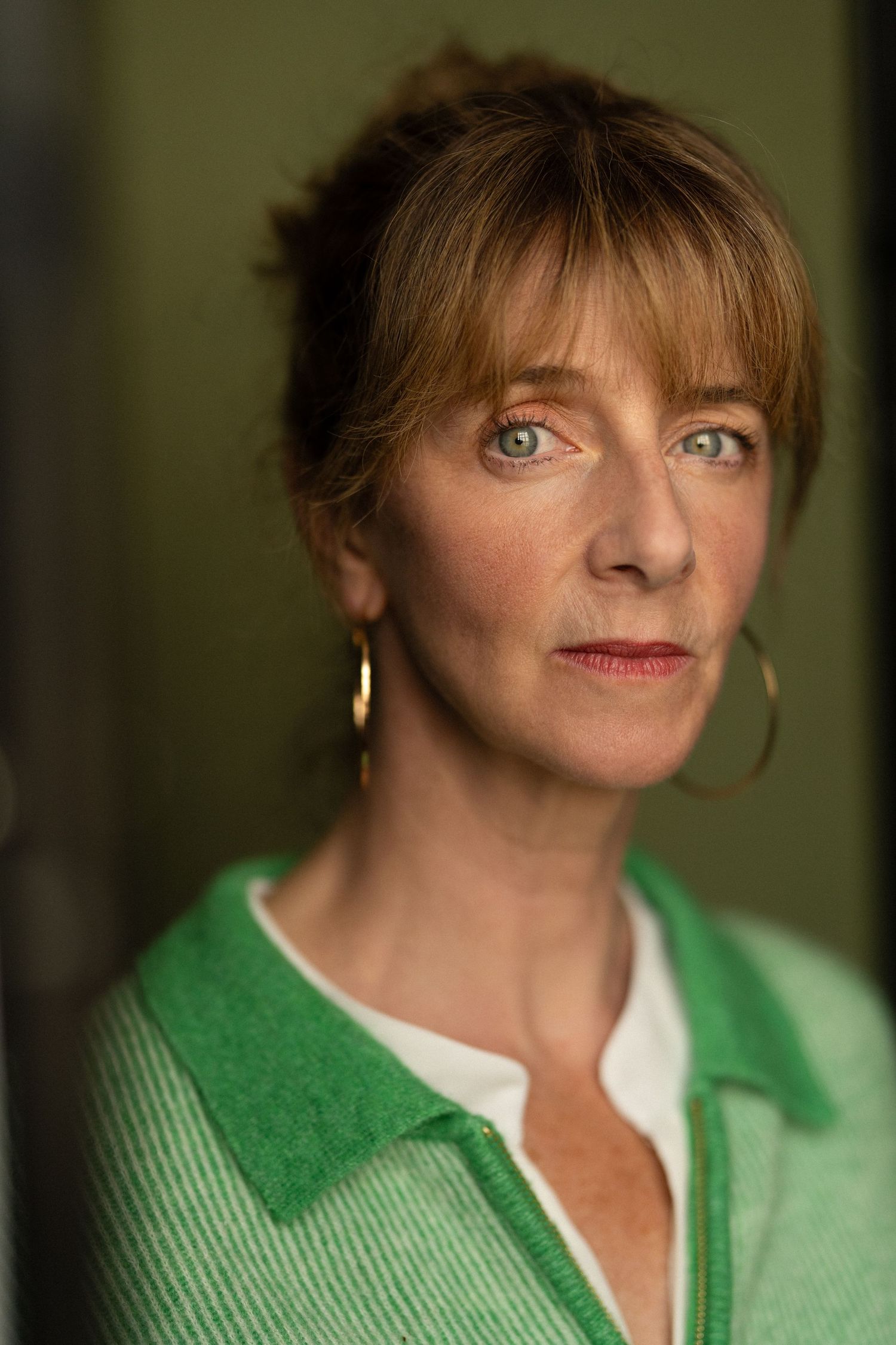Middle-aged woman with auburn hair and green eyes wearing gold earrings and a green cardigan.