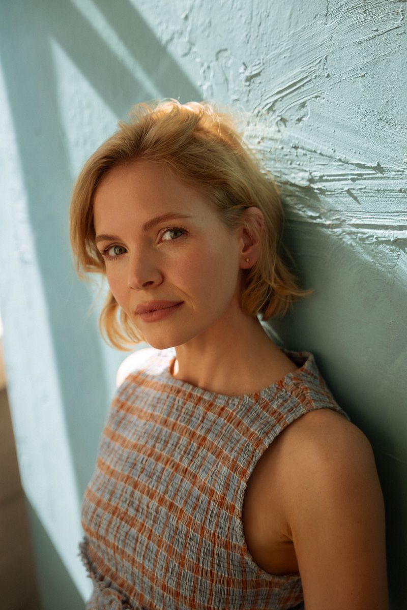 Blonde woman in plaid sleeveless top leaning against a light blue textured wall in warm sunlight.