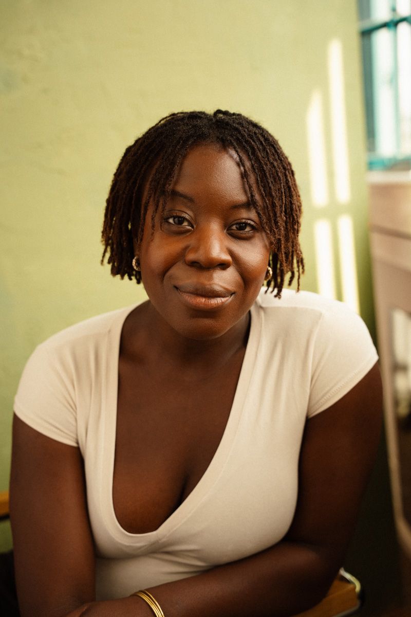 Young woman with braided hair smiling warmly, wearing a white top, seated against a green wall.