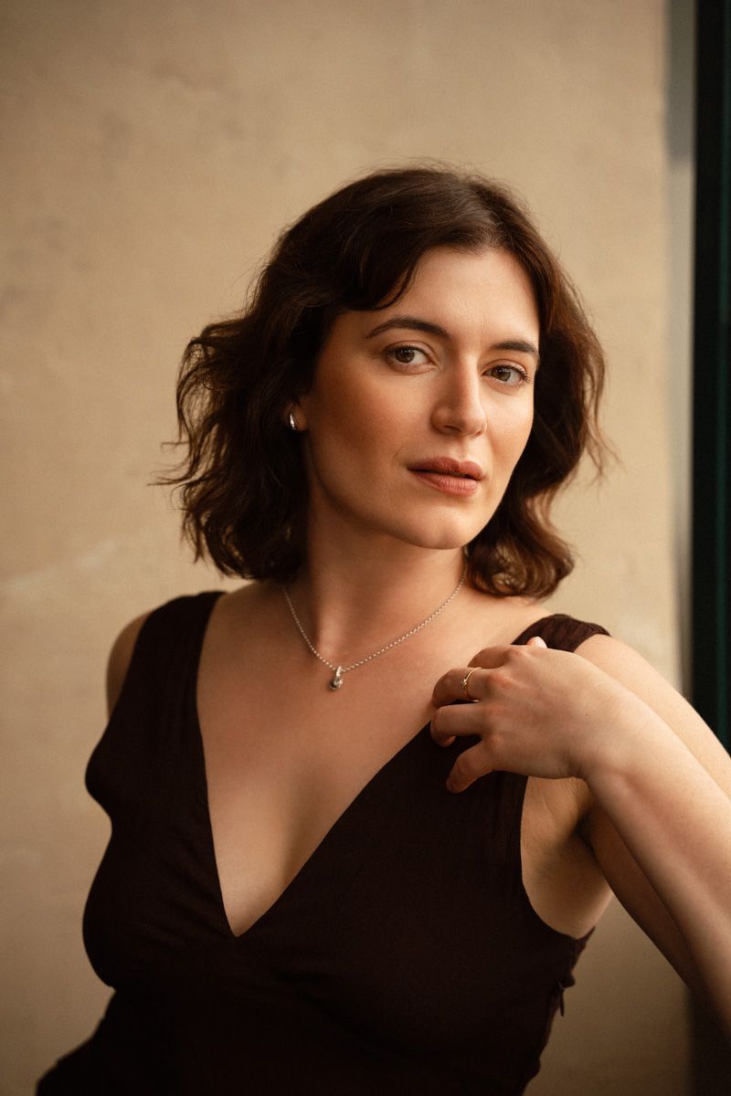 A young woman with short wavy dark hair wearing a brown v-neck dress and silver necklace poses gracefully indoors.
