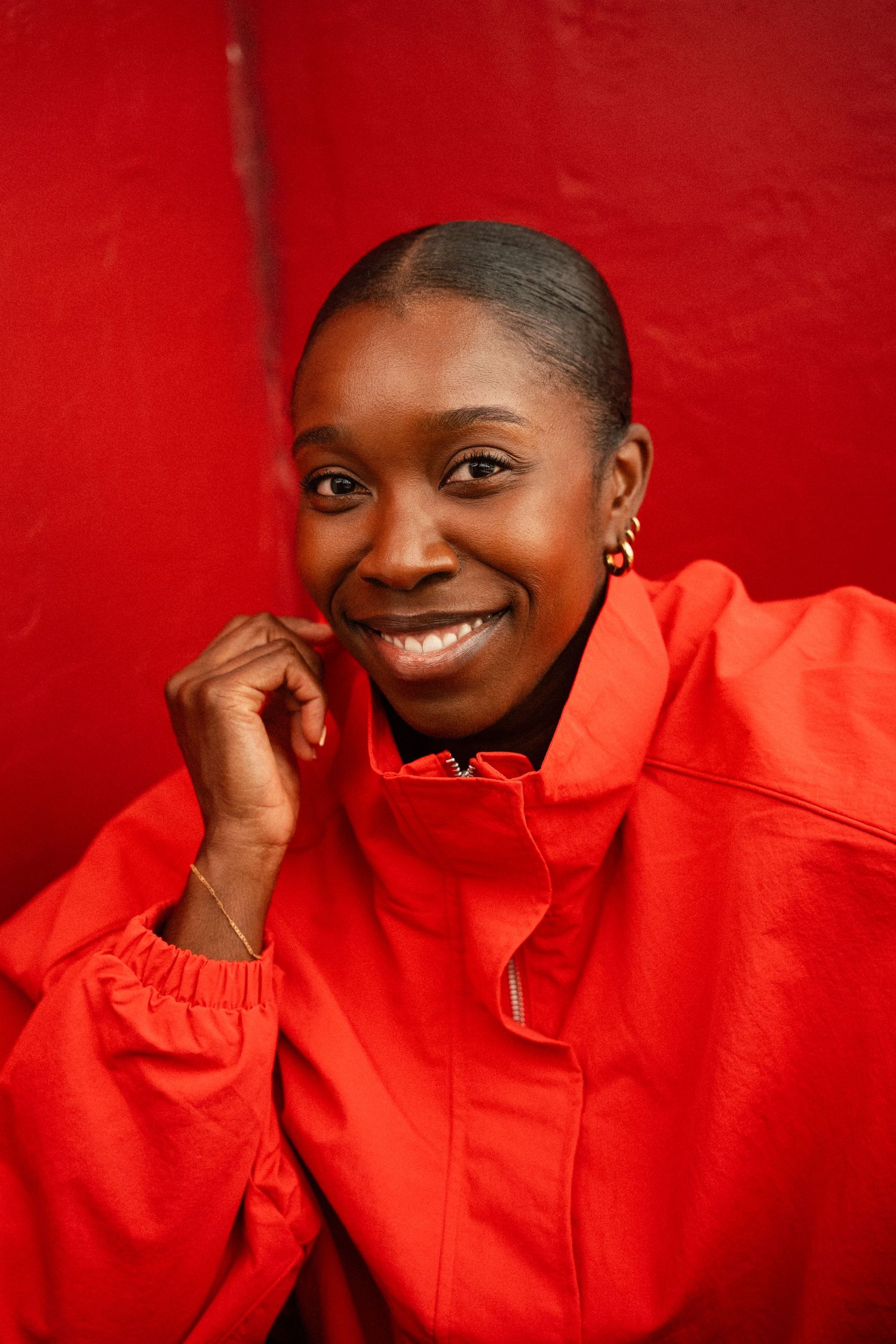 Smiling young woman in a red jacket posing against a red wall with hand raised to chin.