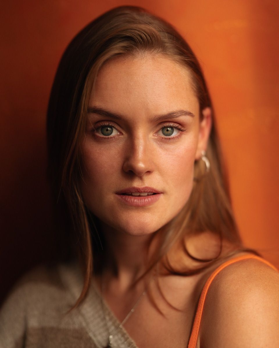 Close-up portrait of a young woman with green eyes, freckles, and long auburn hair against an orange background.
