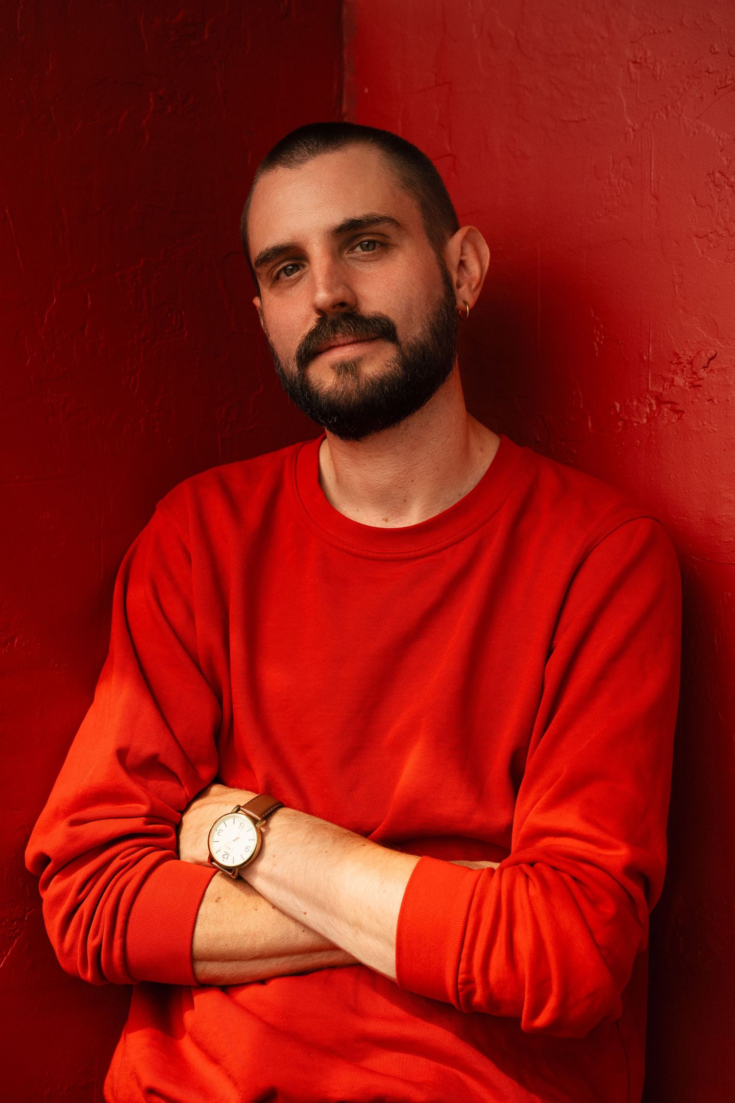Bearded man in red sweatshirt with arms crossed, wearing a watch, leaning against a red wall.