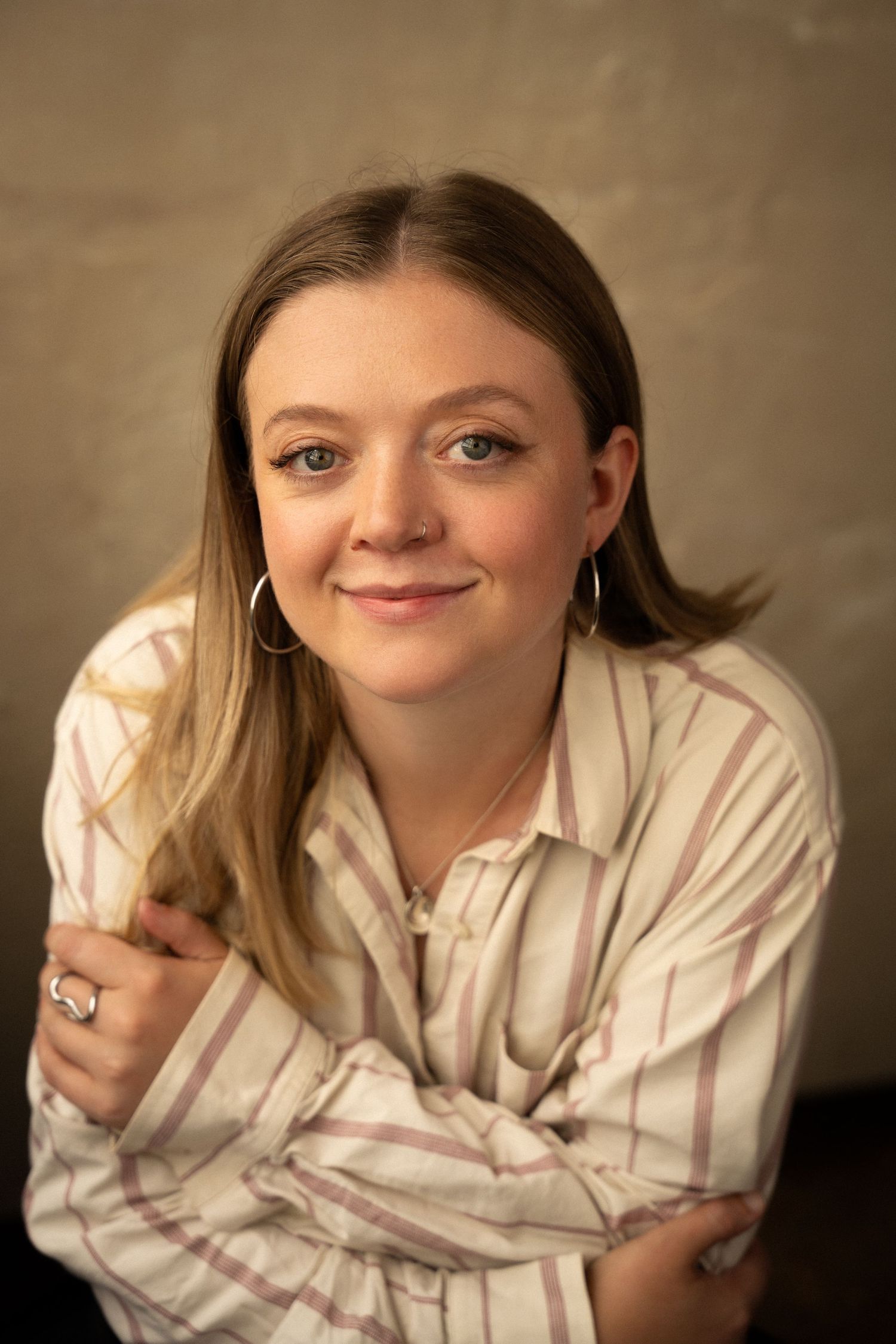 Young woman with blonde highlights smiling warmly, wearing a striped shirt and hoop earrings in a portrait photo.
