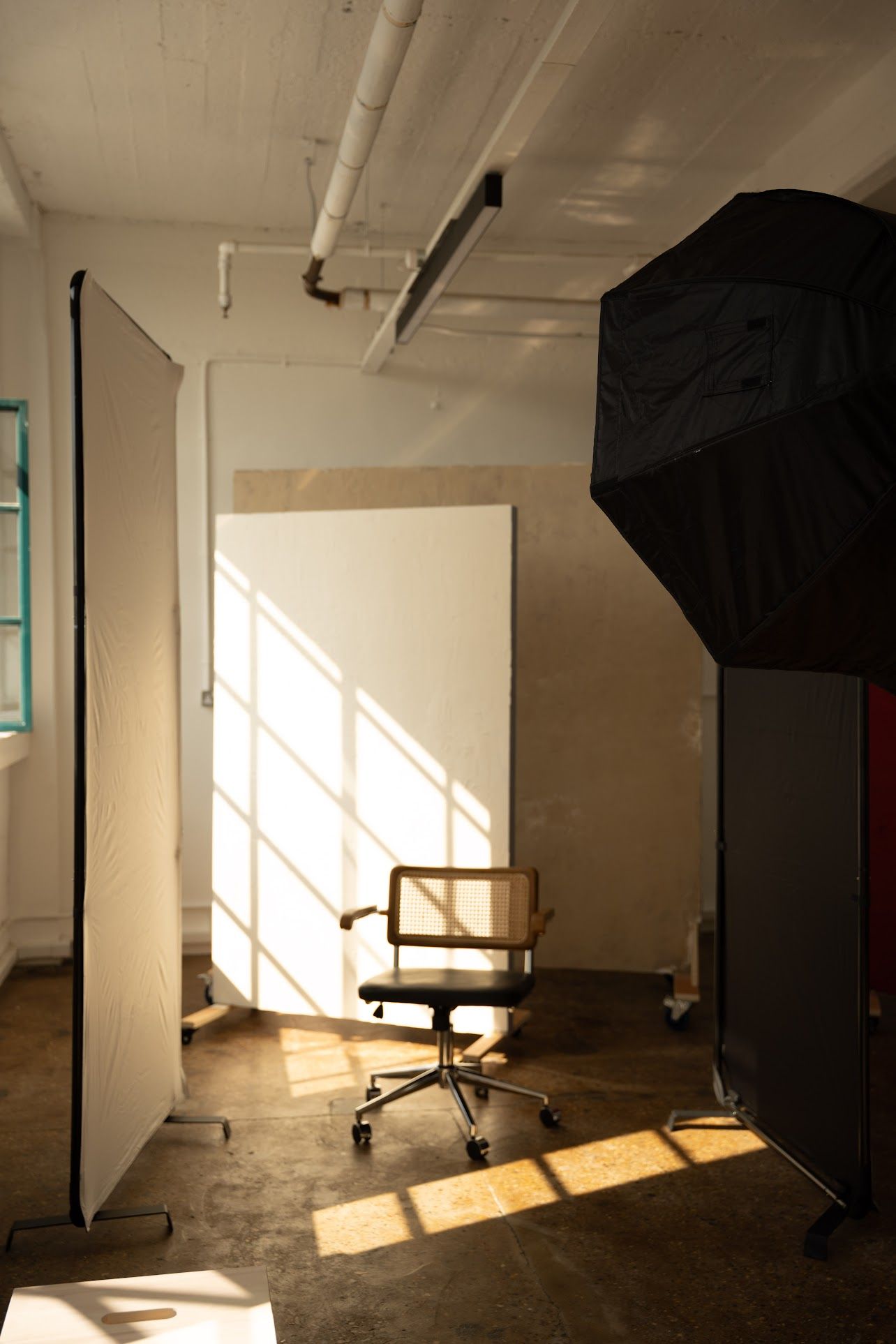 A professional photography studio setup with a white backdrop, office chair, softbox light, and natural sunlight streaming through windows.