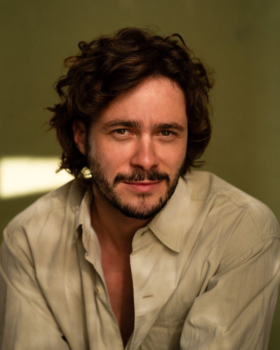 Young man with curly dark hair and beard, wearing an open white shirt, smiling warmly against a green background.