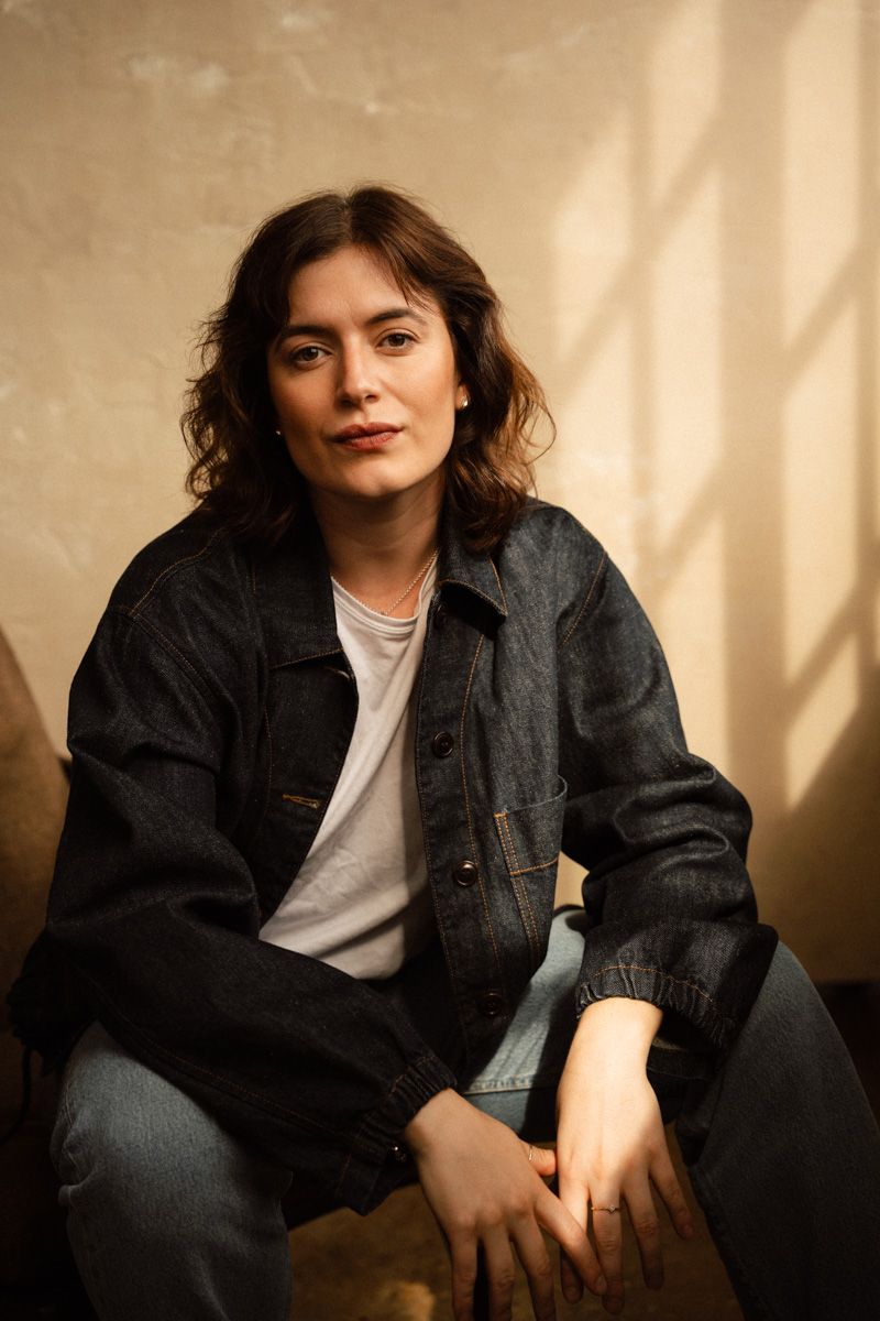 Young woman in dark denim jacket and jeans sitting relaxed against a warm beige wall with natural light.