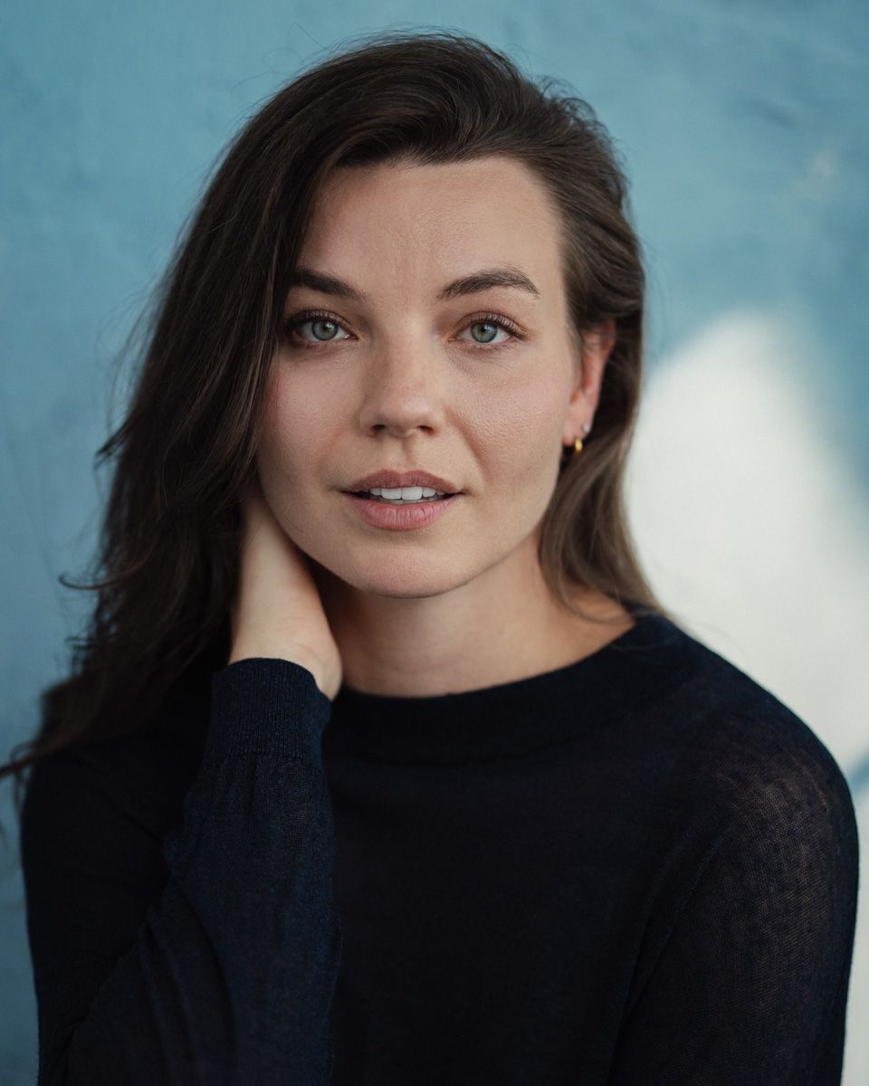 Young woman with dark hair in black sweater poses confidently against a blue background in a professional headshot.