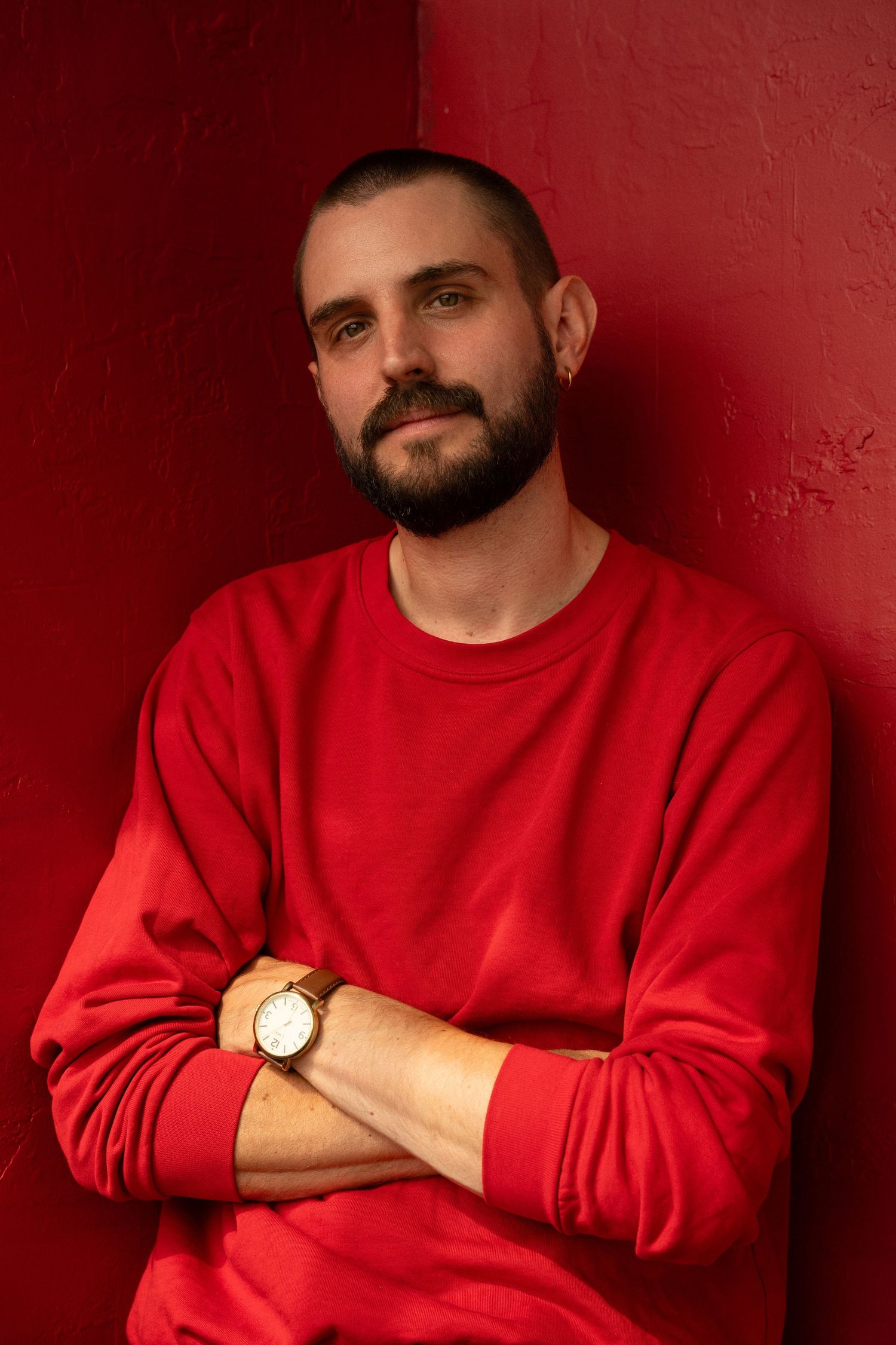 Bearded man in red sweatshirt with crossed arms and gold watch leaning against a red wall.