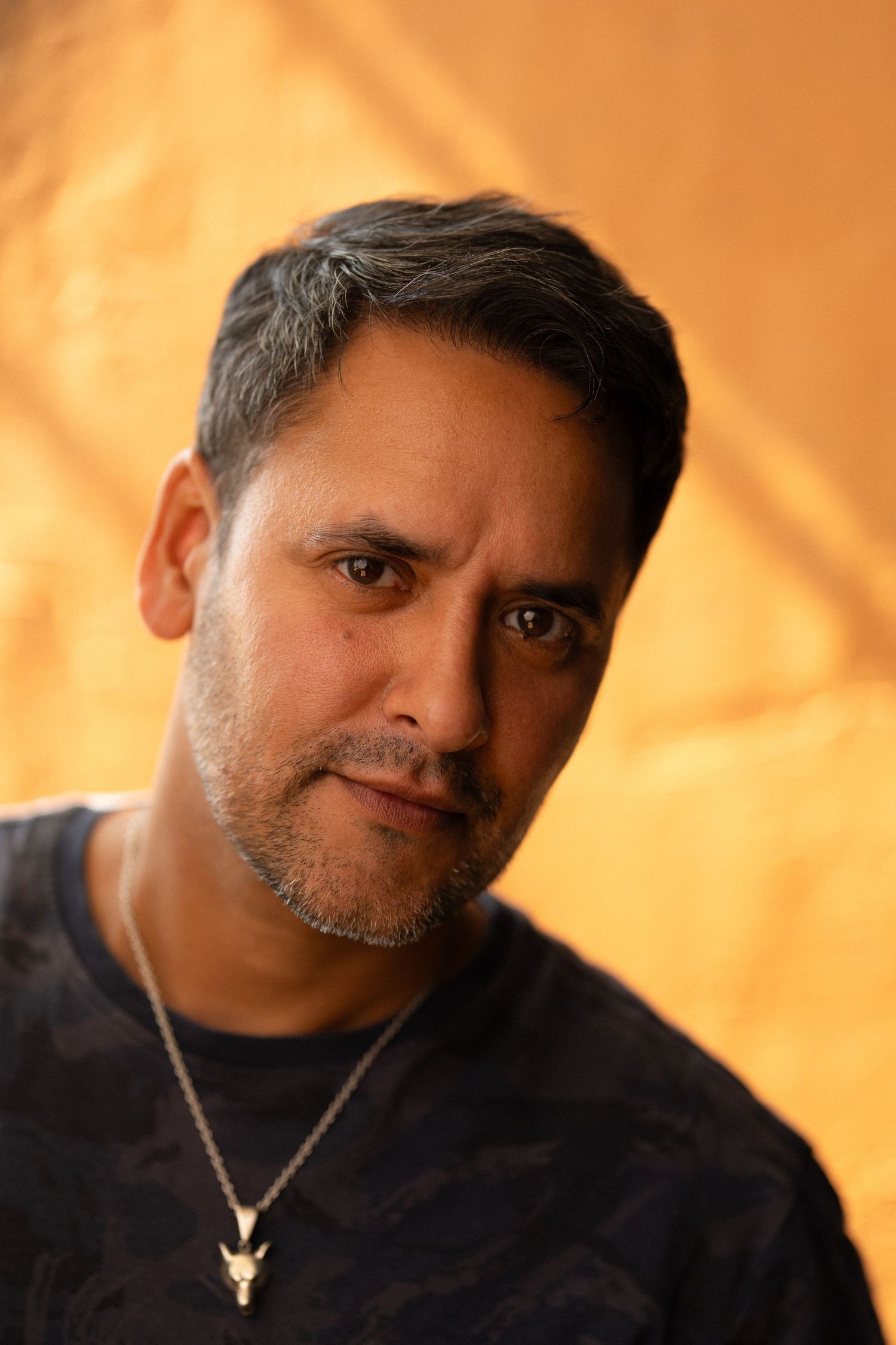 Middle-aged man with salt-and-pepper hair, stubble, and a necklace poses against a golden yellow fabric background.