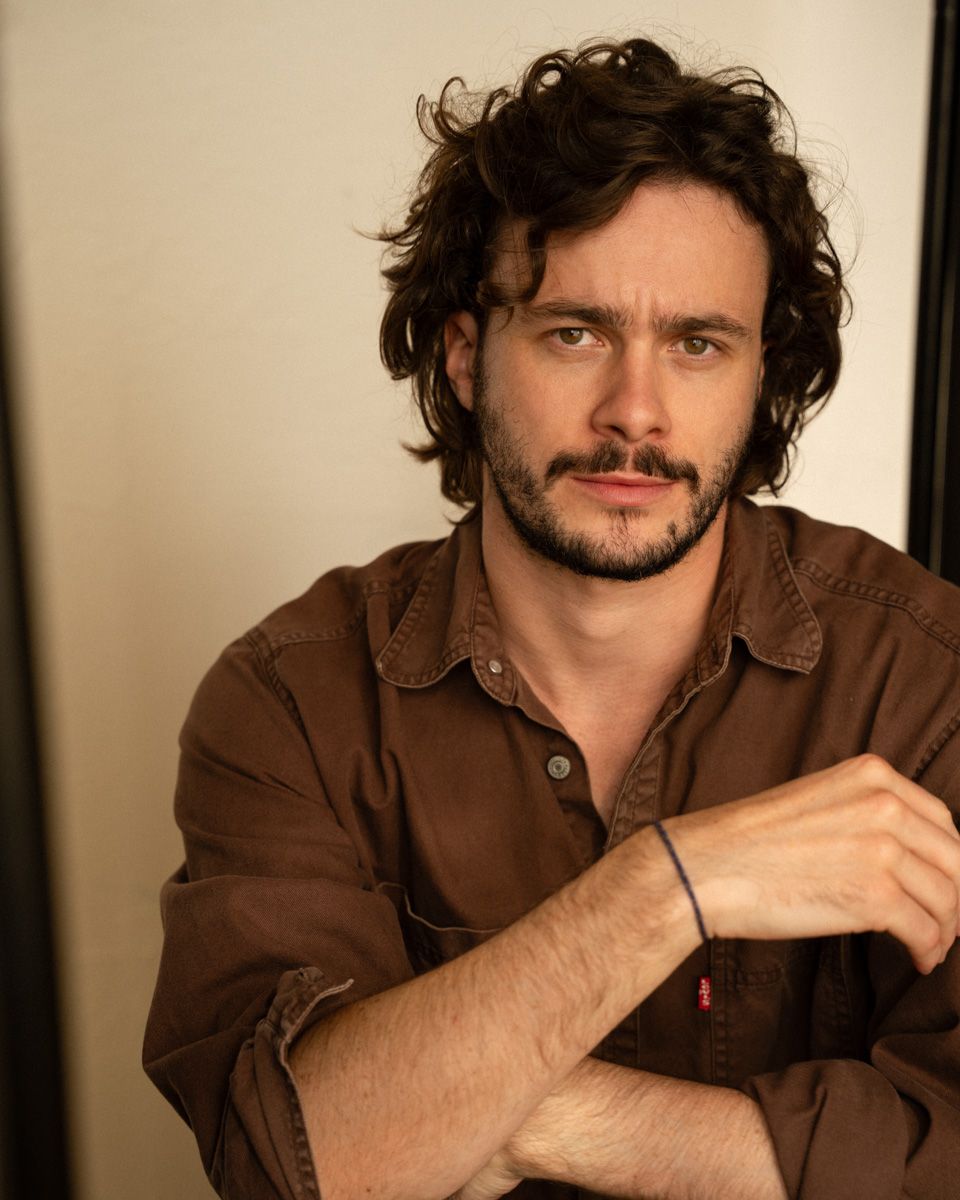 Young man with curly dark hair and beard wearing a brown shirt, arms crossed, posing for a portrait photo.