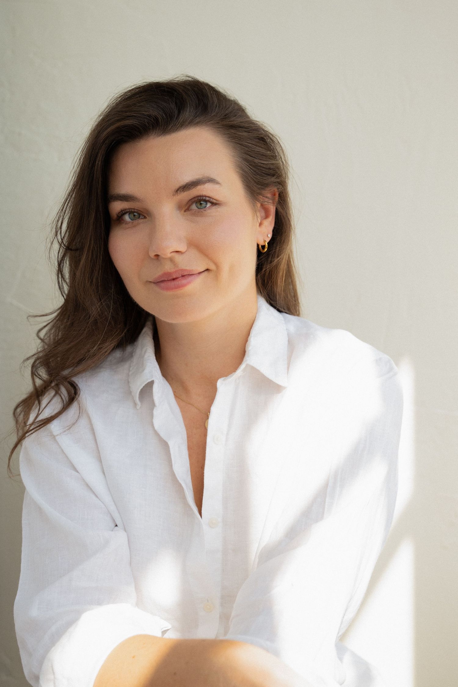 Young woman with brown hair wearing a white linen shirt, smiling softly against a light background.