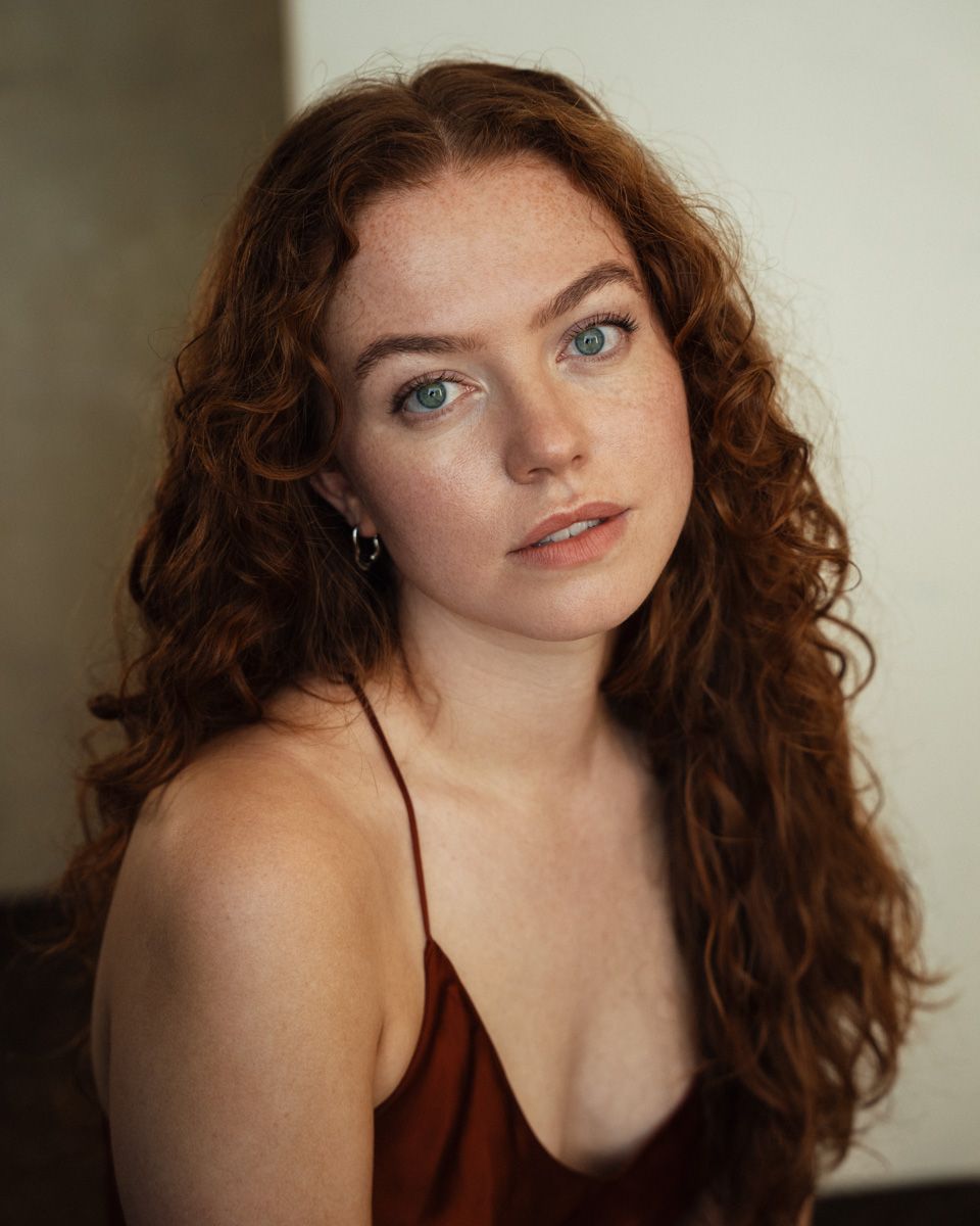 Young woman with curly red hair, freckles, and green eyes wearing a rust-colored top in a portrait photo.